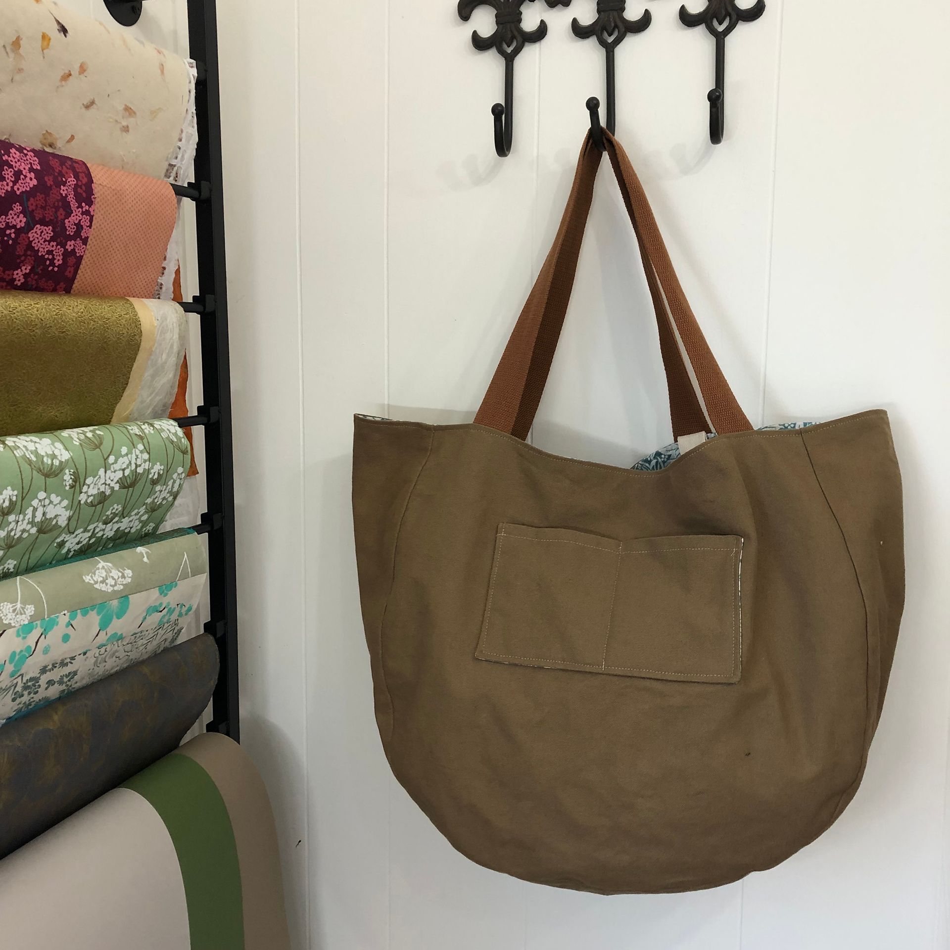 Brown tote bag hanging on a white wall with black hooks. Stacks of fabric are visible on the left.