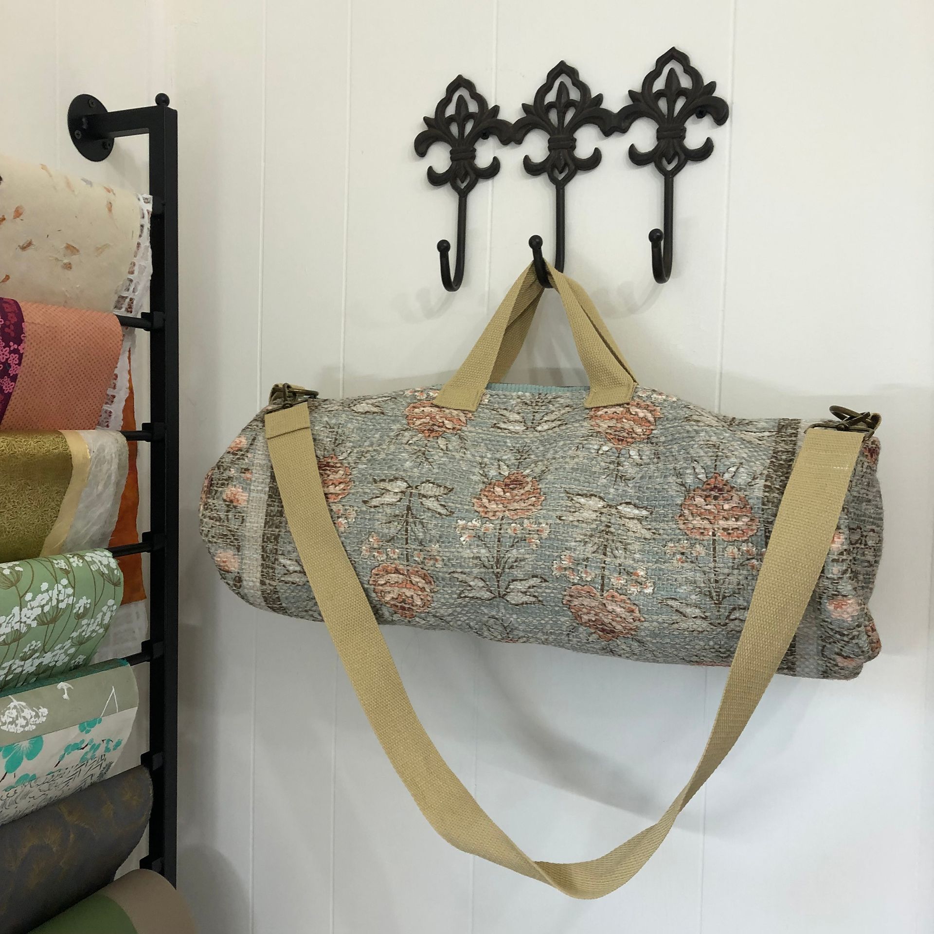 Fabric bag with floral print hanging on a wall with decorative hooks, next to fabric rolls.