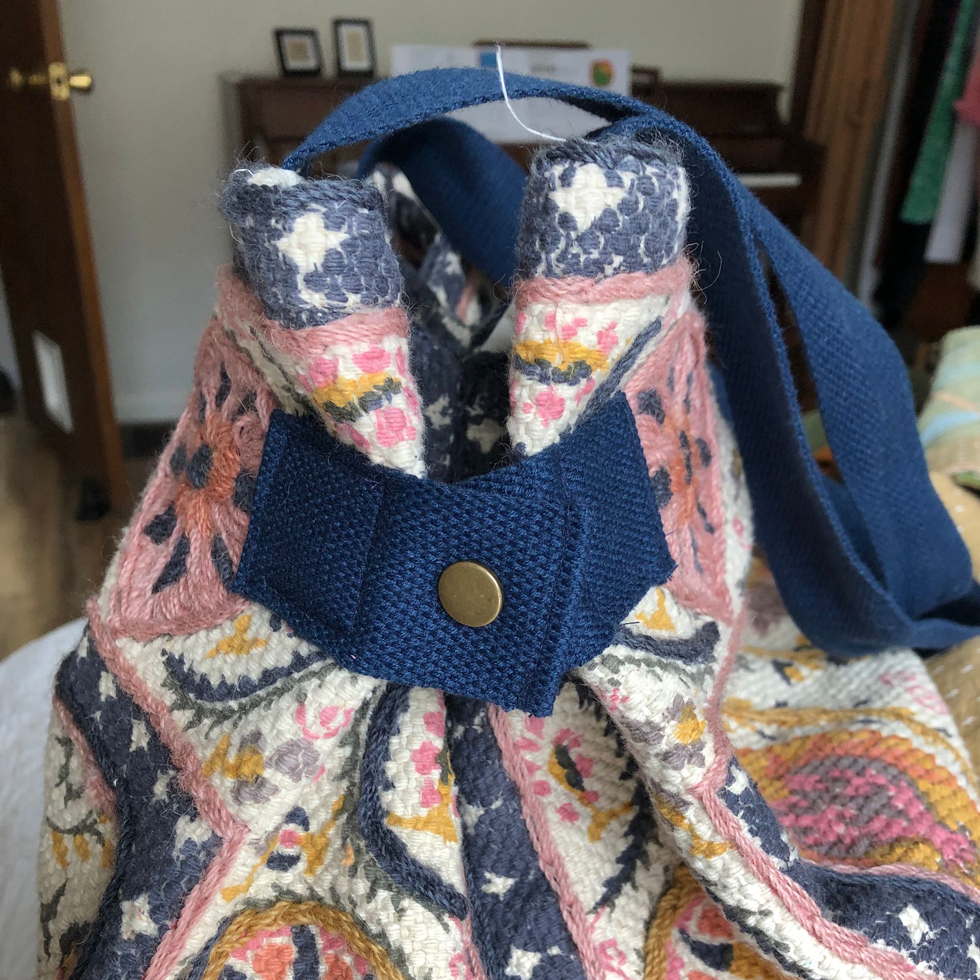 Cream tote bag with floral design, dark blue straps, hanging on a wall next to fabric rolls.