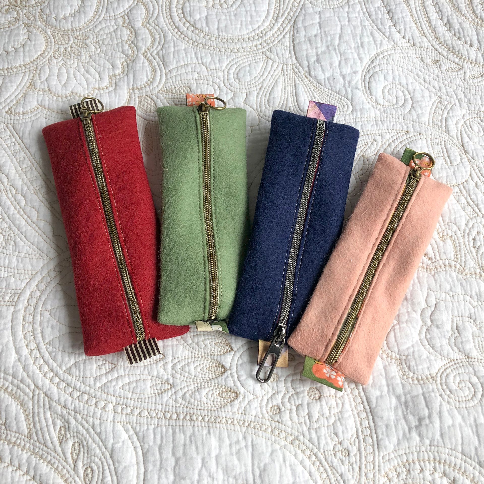 Four rectangular pouches: red, green, blue, pink, with gold zippers, lying on a white quilted surface.