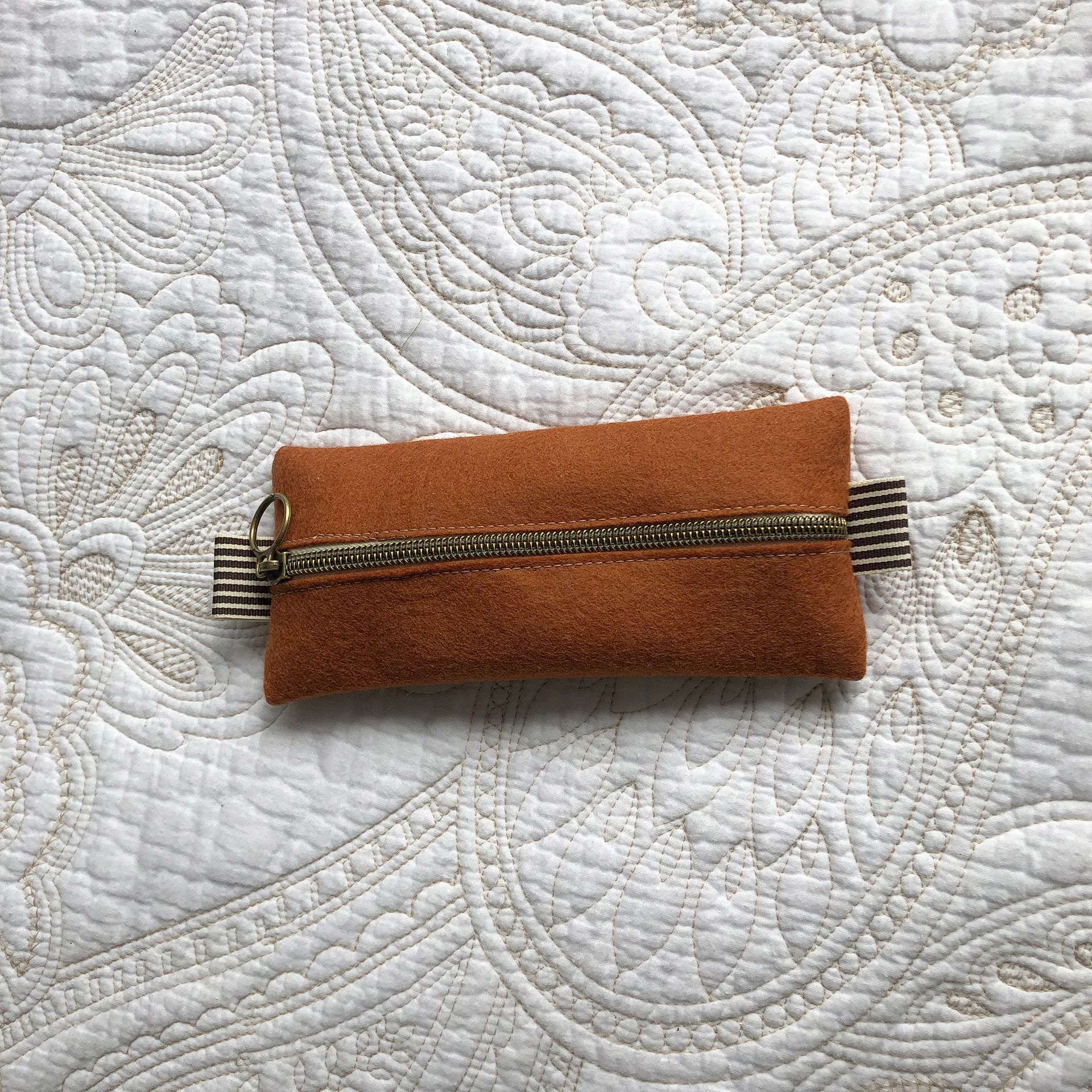 Small, brown zippered pouch with gold and striped detailing, placed on a white paisley patterned blanket.