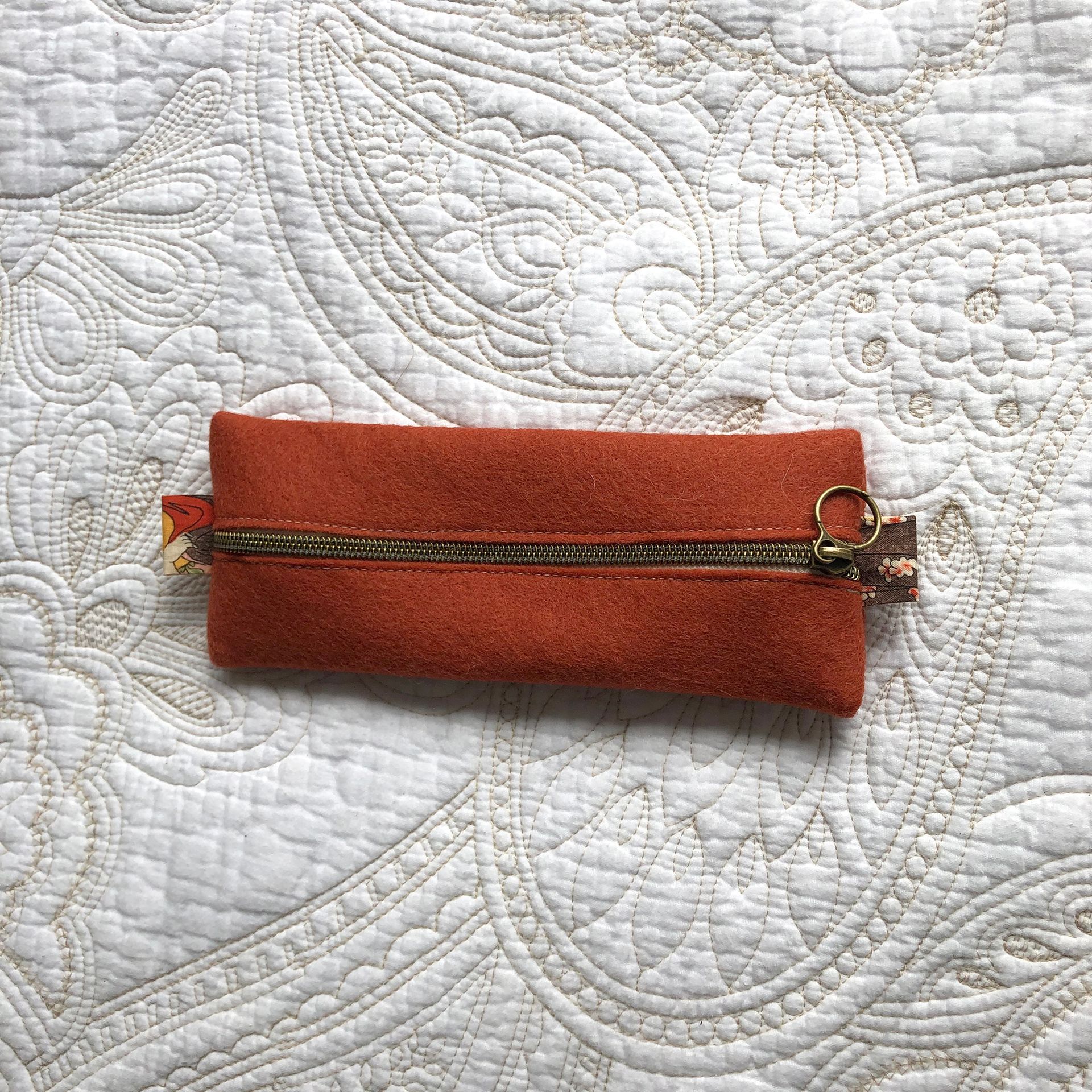 Small rust-colored suede zippered pouch on a white paisley patterned surface.