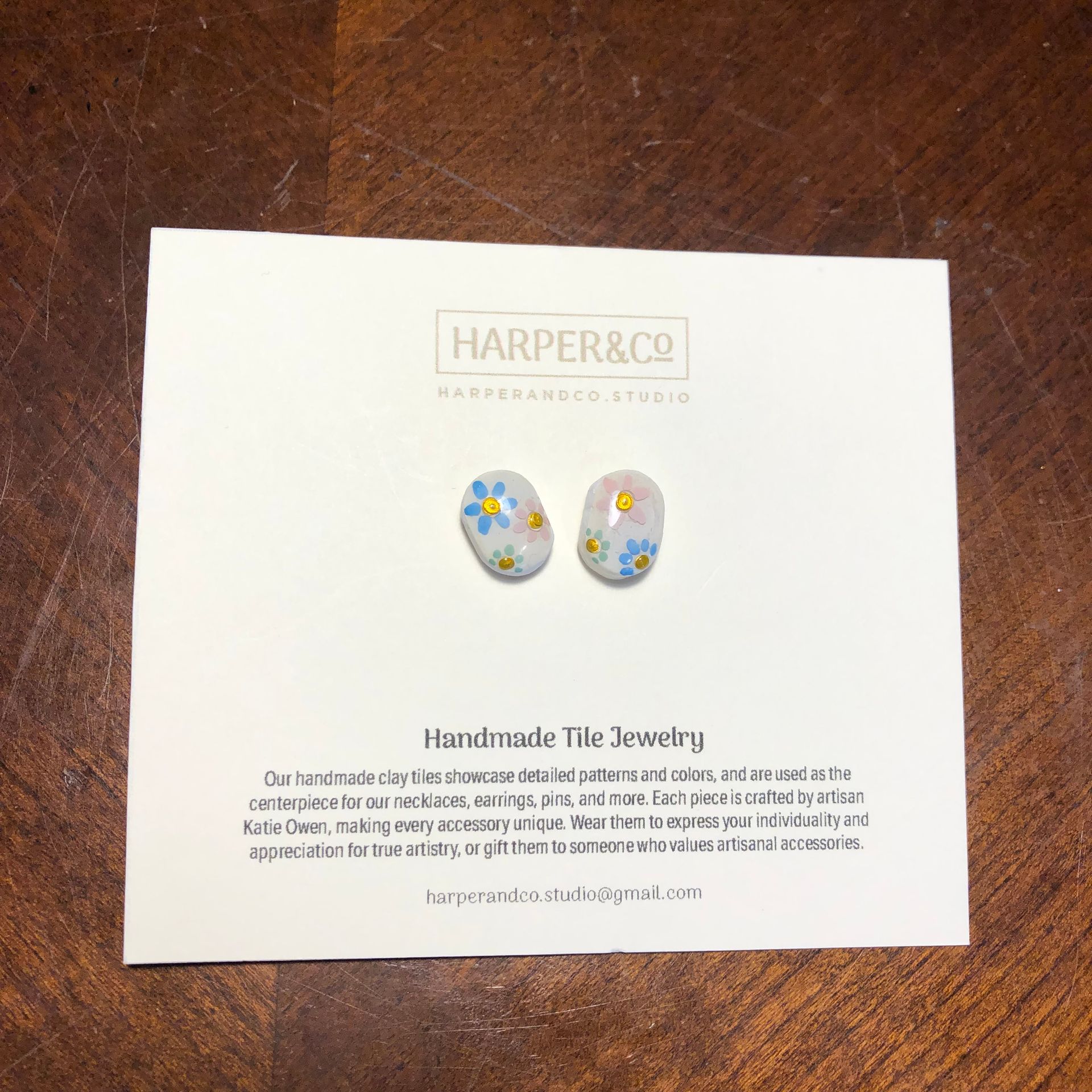 Earrings with blue and yellow flowers on a white card with text, on a wooden surface.