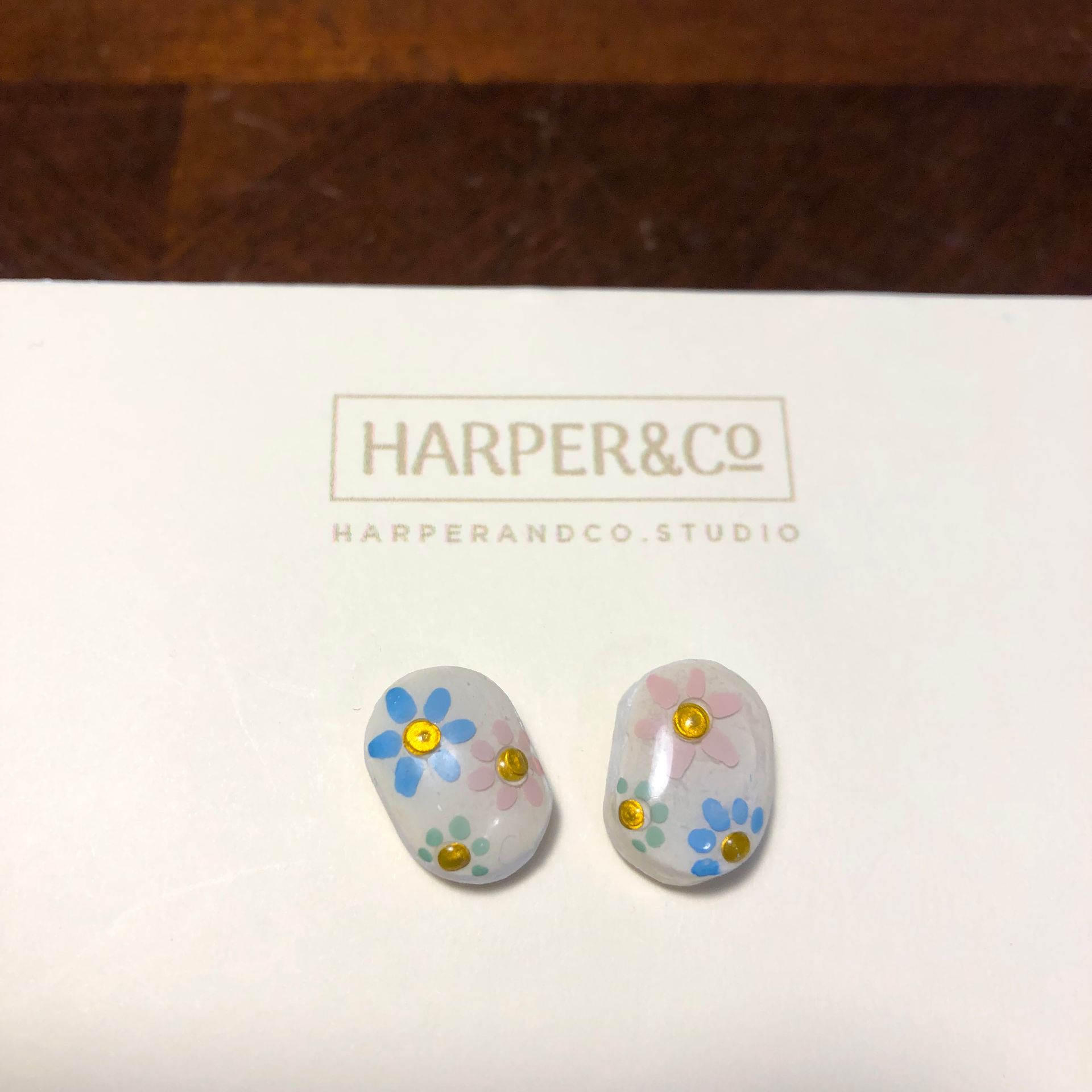 Earrings with blue and yellow flowers on a white card with text, on a wooden surface.