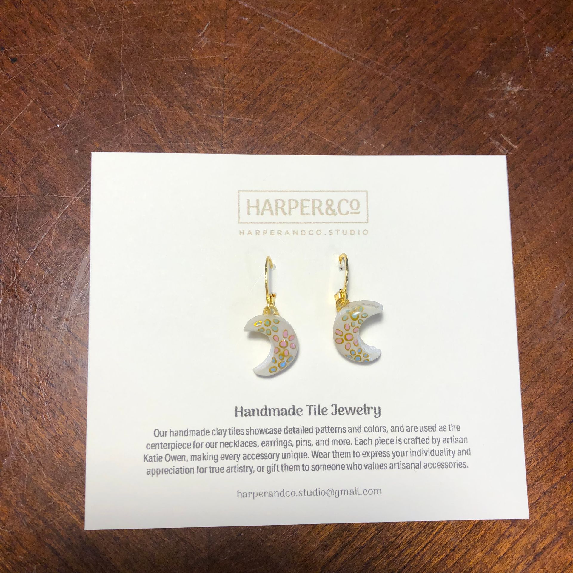 Earrings displayed on a card; white crescent-shaped tiles with floral designs, gold accents, on a wooden surface.