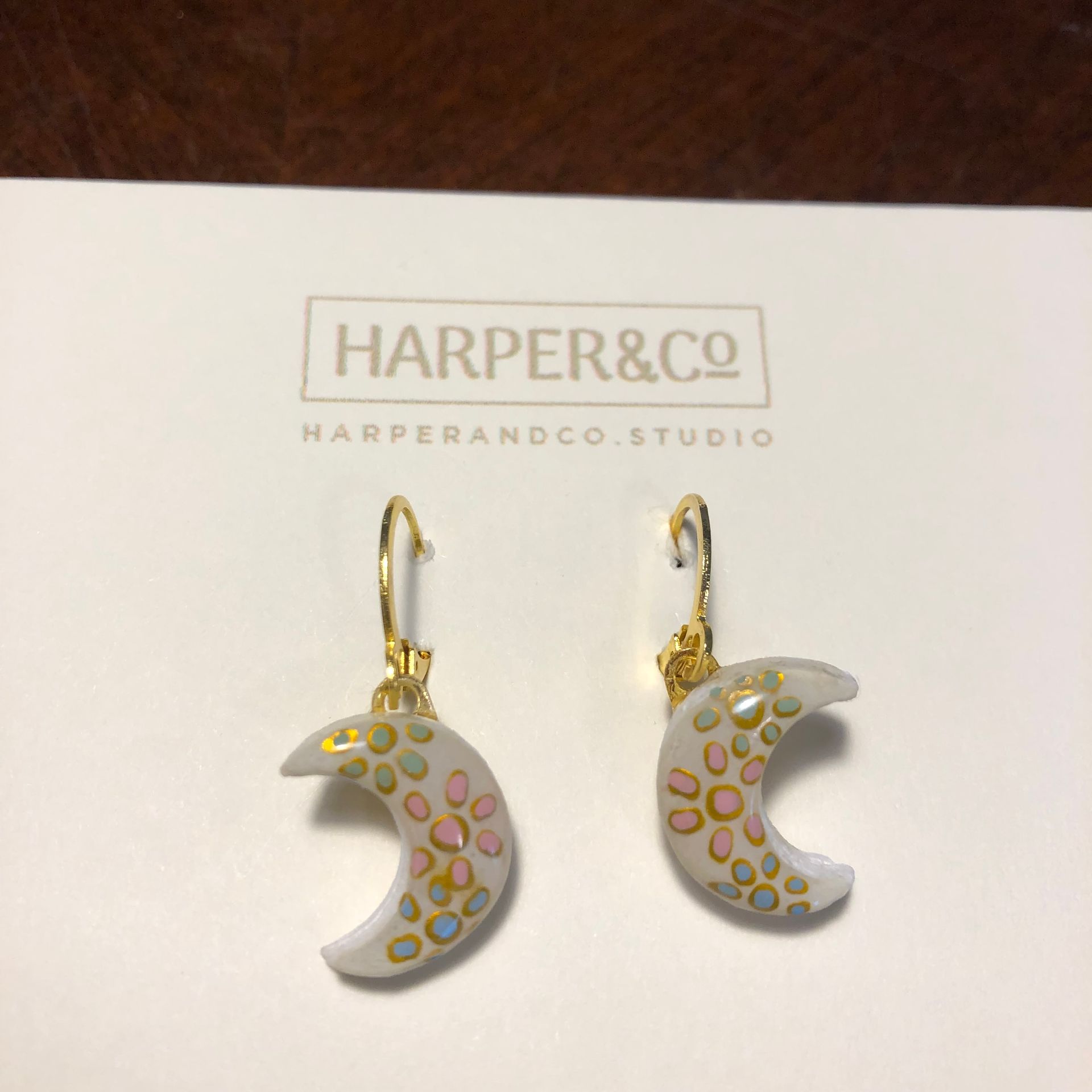 Earrings displayed on a card; white crescent-shaped tiles with floral designs, gold accents, on a wooden surface.