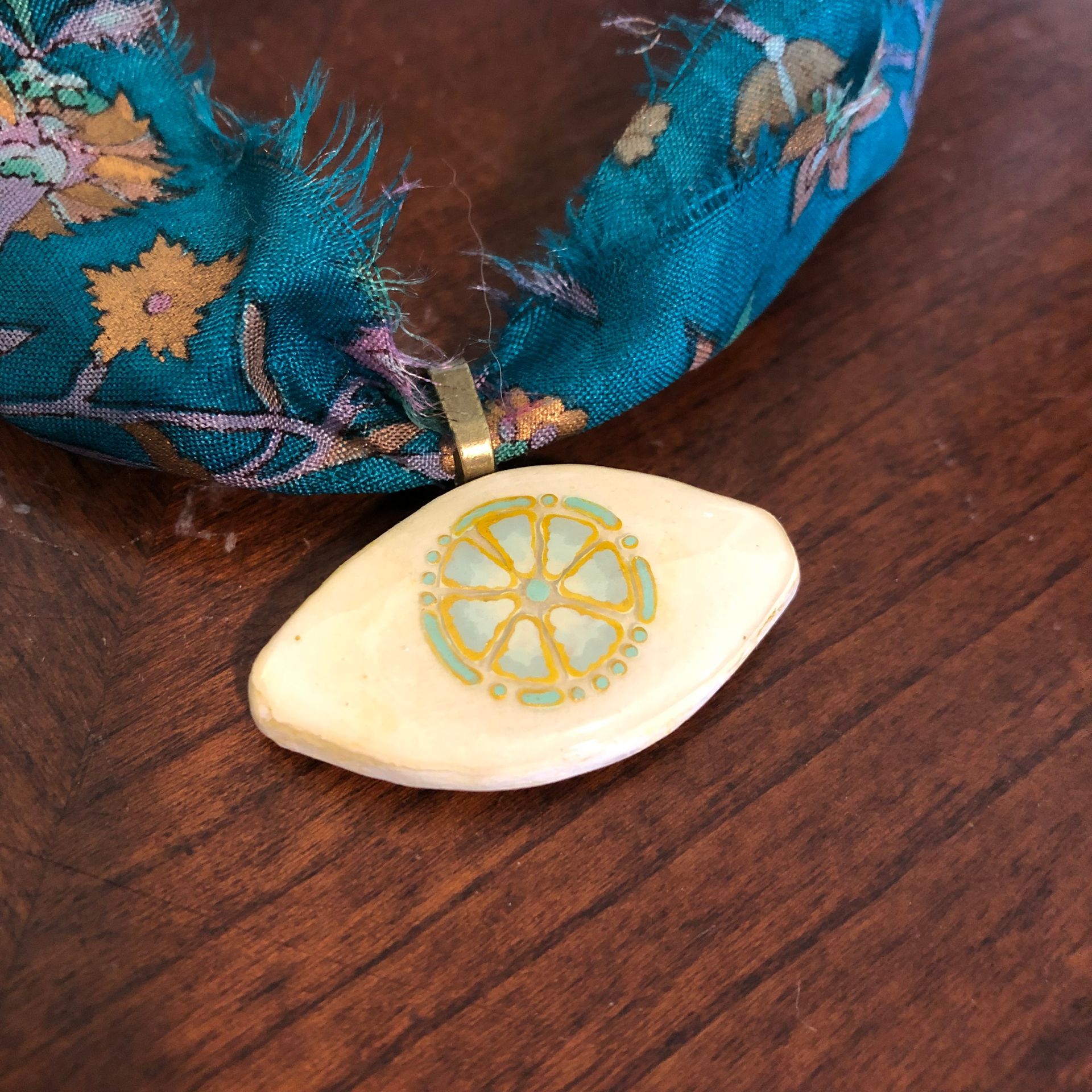 Teal silk scarf necklace with floral print, gold chain clasp, and ivory eye pendant with gold detail.