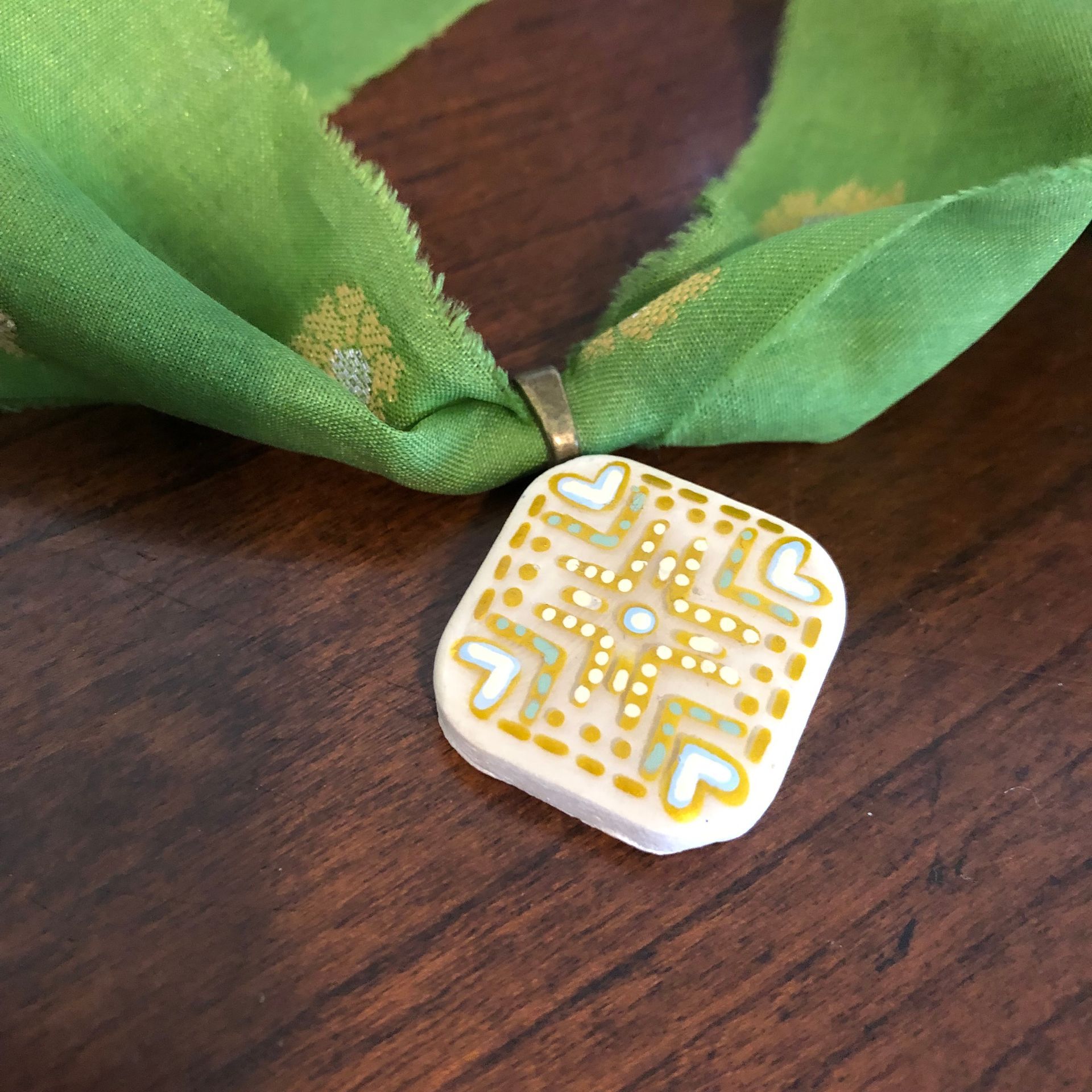 Green scarf necklace with gold diamond pattern and square pendant on wooden surface.