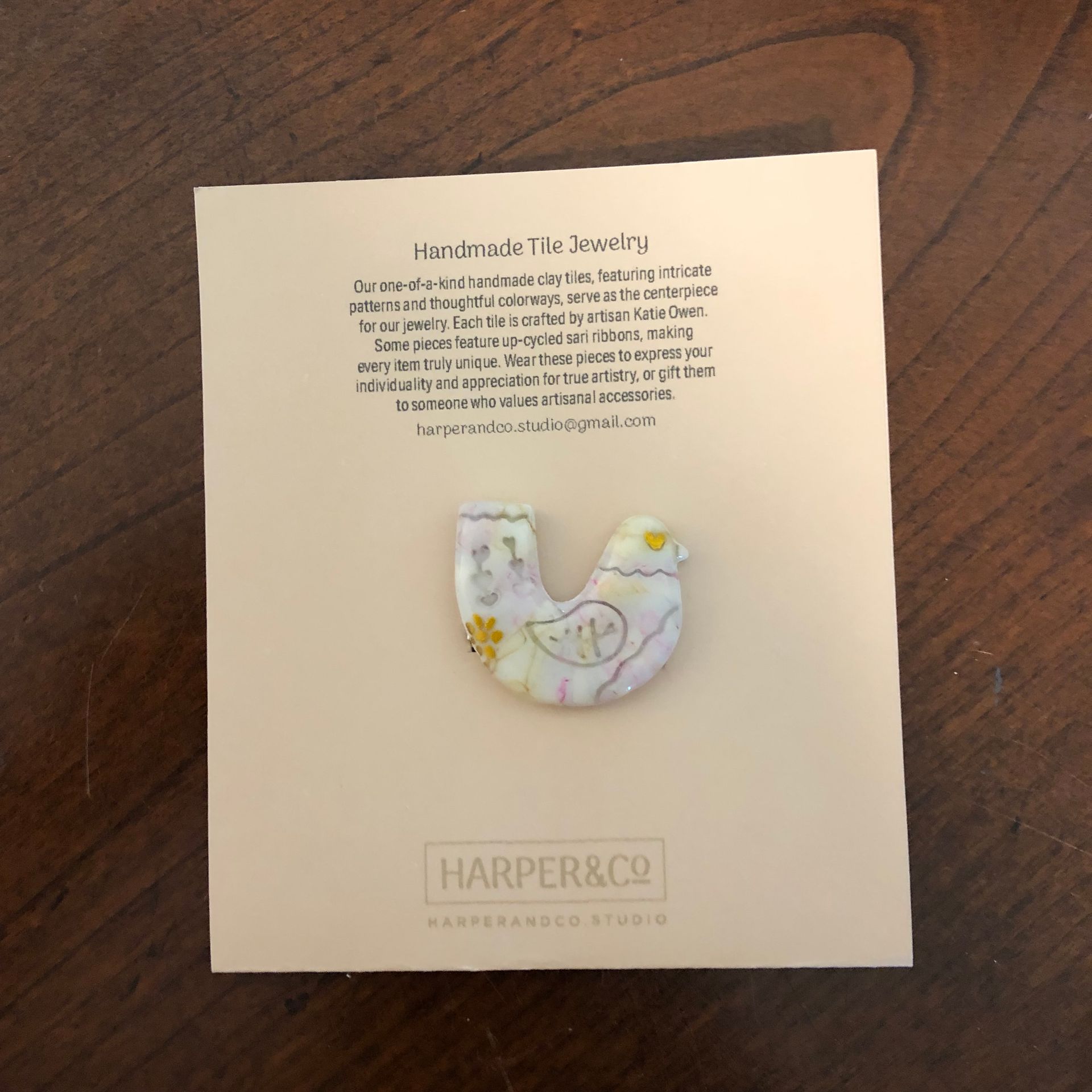 Bird-shaped handmade tile jewelry on a cream-colored card with text, on a wood surface.