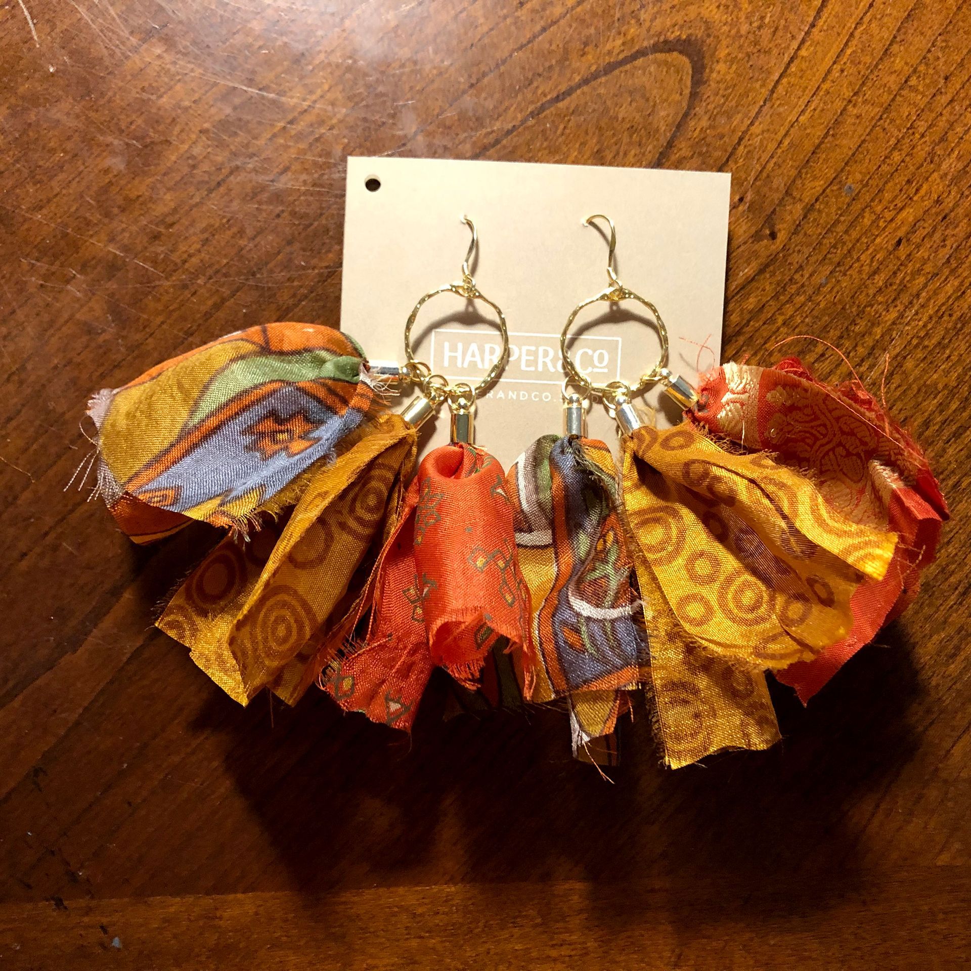 Gold hoop earrings with colorful fabric tassels, on a wooden surface, with a tag.