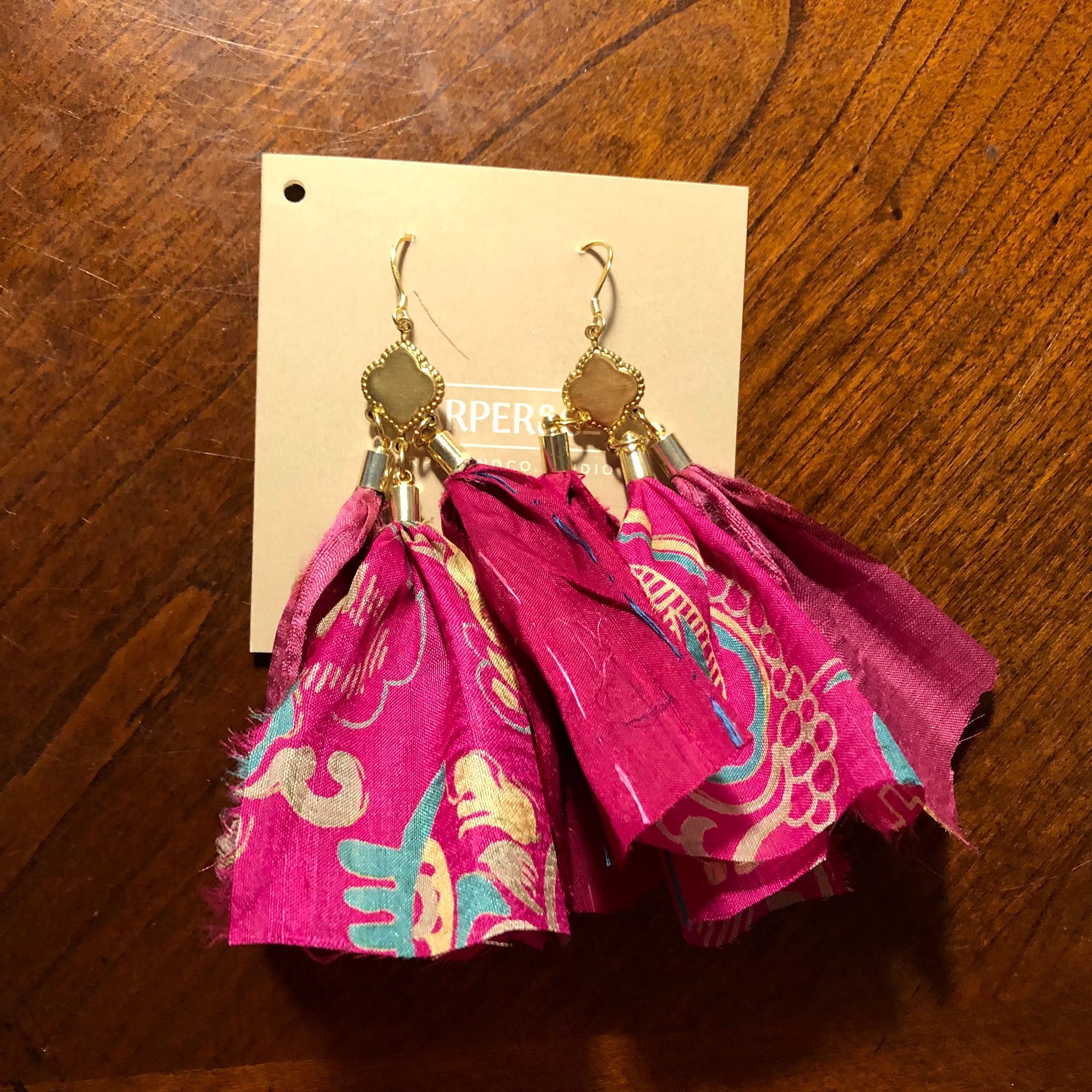 Gold hoop earrings with colorful fabric tassels, on a wooden surface, with a tag.