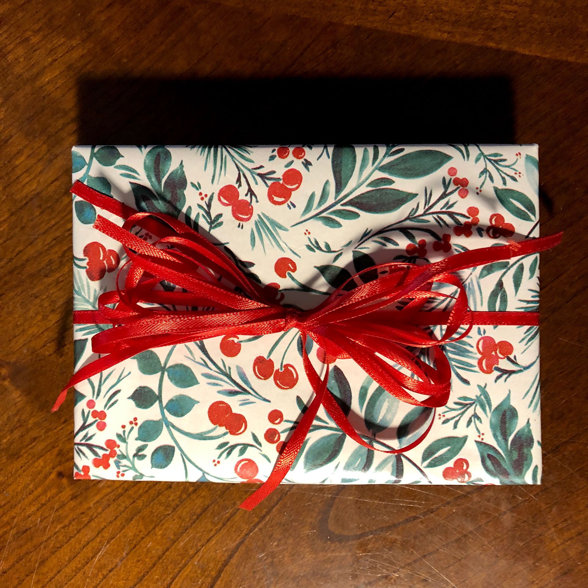 Gift wrapped in white paper with red berries and green leaves, tied with a red ribbon on a wooden surface.