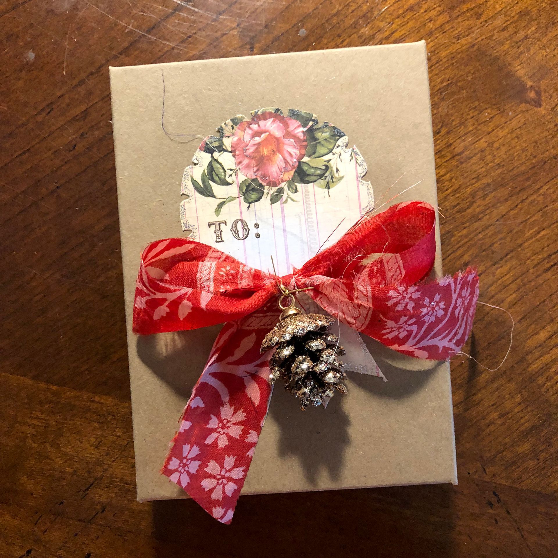Gift box with red ribbon and pinecone charm. Floral sticker, on a wooden surface.