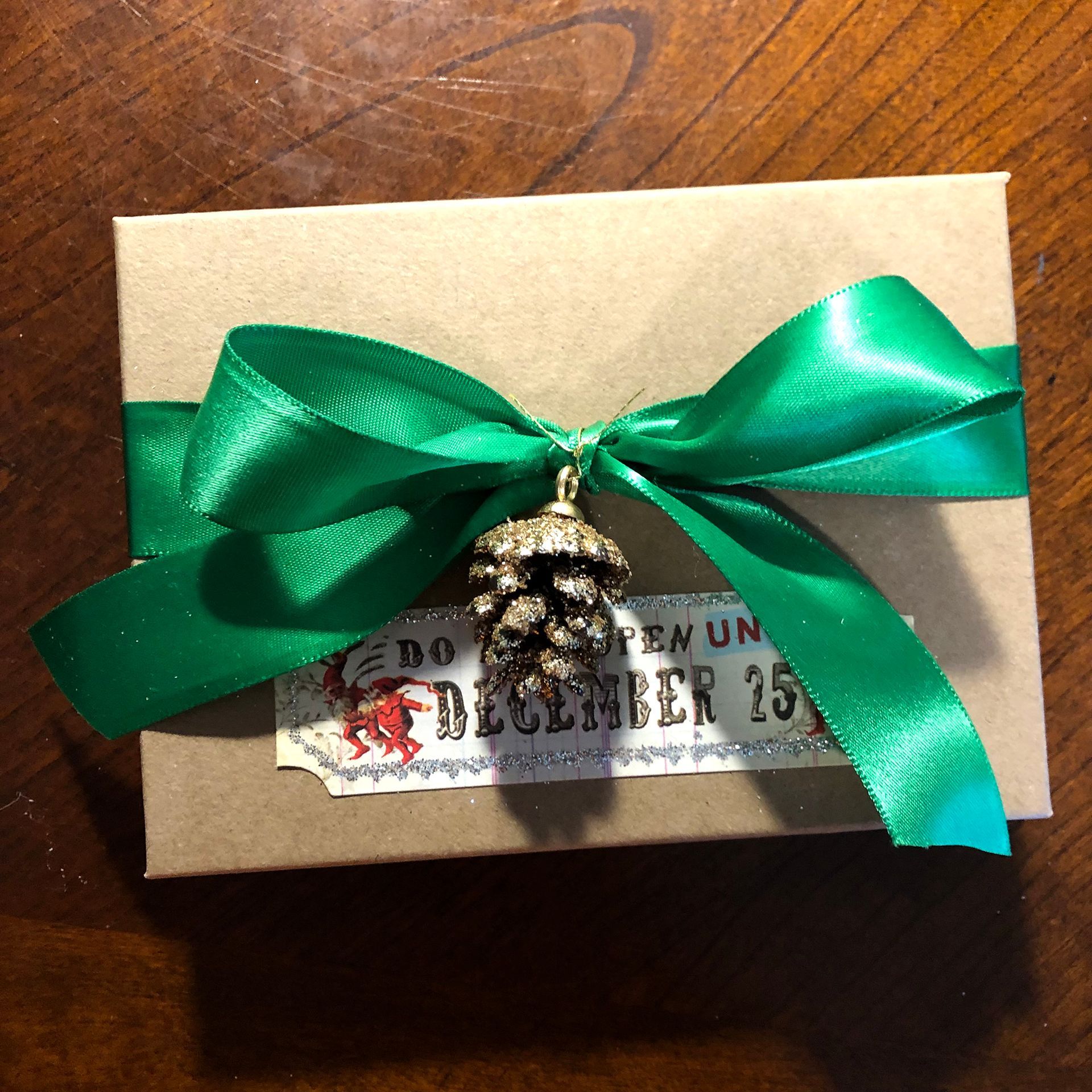 Gift wrapped in brown paper with green satin ribbon and gold pinecone charm; tag reads 