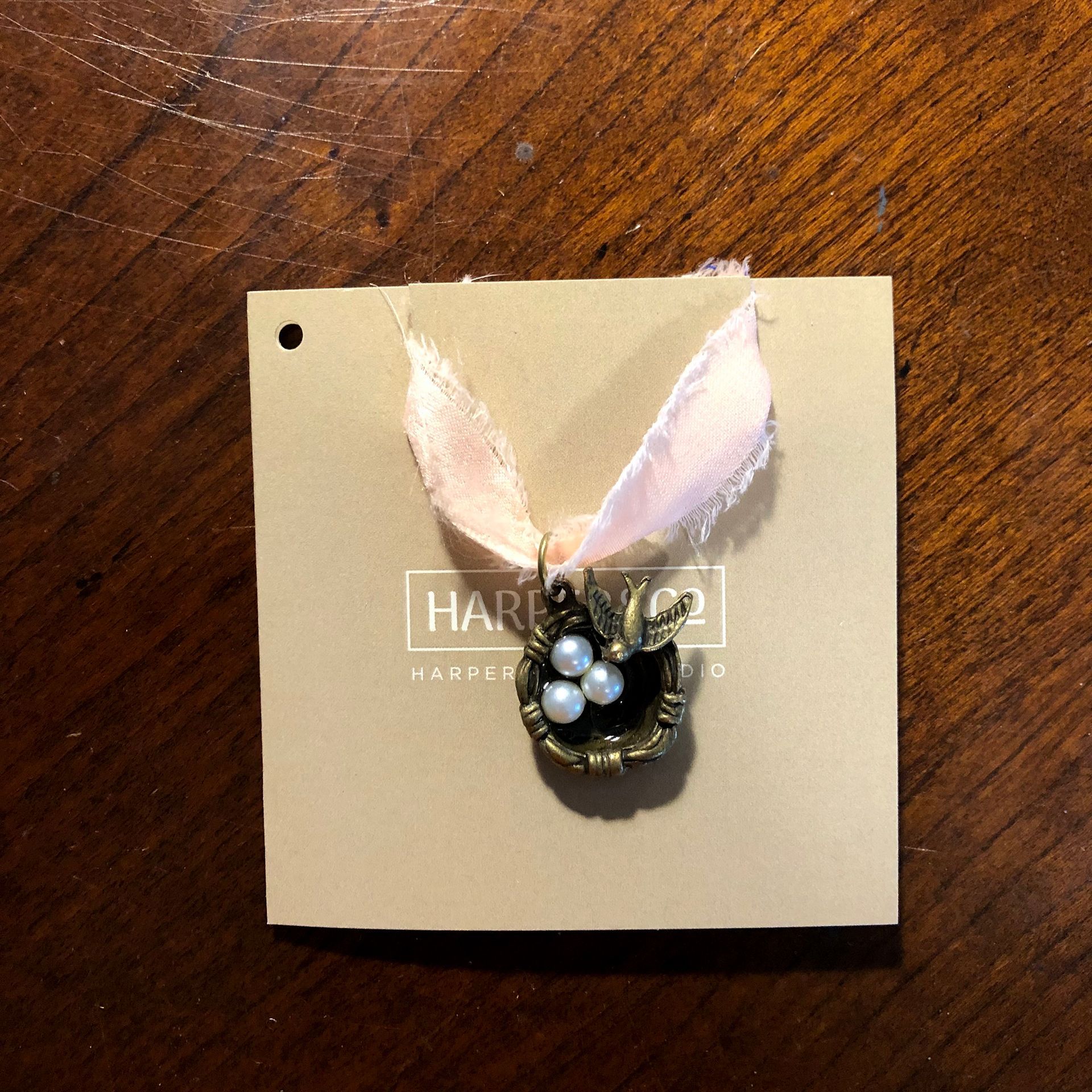 Pendant with three pearls in a nest-like setting, attached to a pink ribbon on a tan tag.