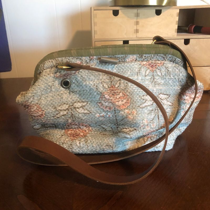 A light blue floral tapestry handbag with a green trim and a brown leather strap, resting on a wooden table.