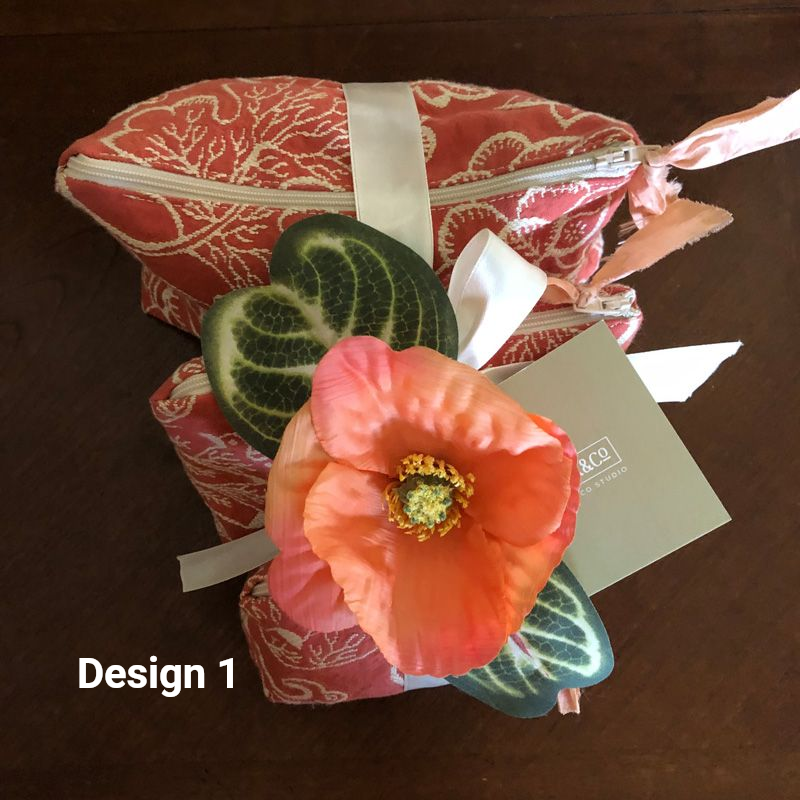 Two coral floral-print fabric pouches stacked together, accented with a large silk poppy, green leaf, and a ribbon.