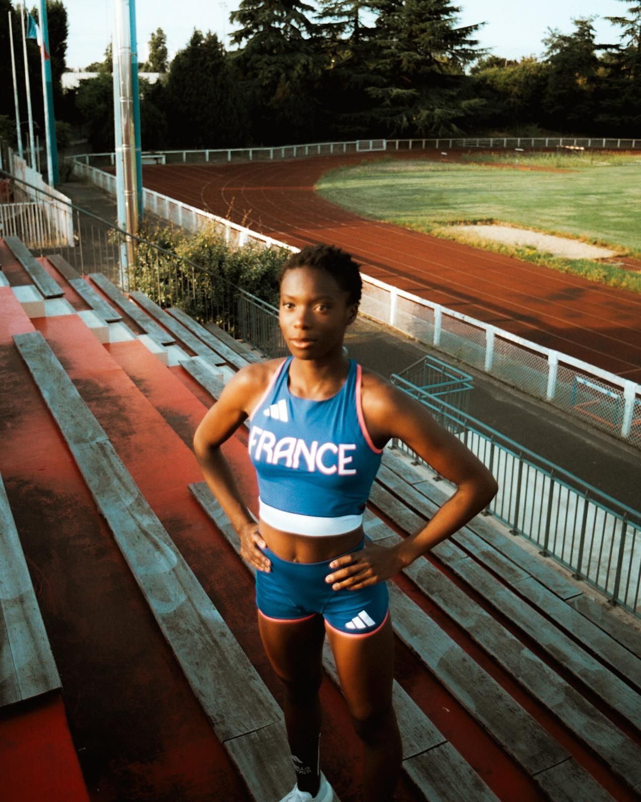 Un athlète en uniforme bleu français se tient debout sur les gradins du stade, les mains sur les hanches.