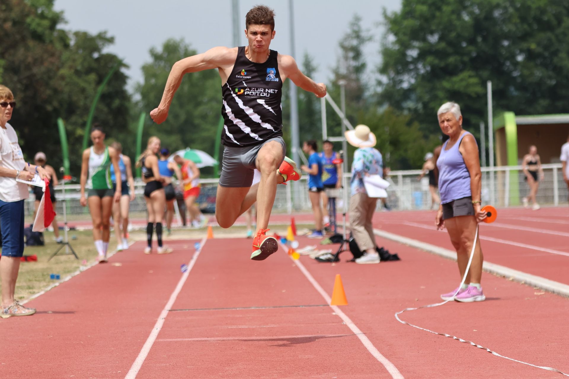 Un athlète triple sauteur en plein vol sur une piste. Piste rouge, herbe verte, journée ensoleillée. Edouard Morin.