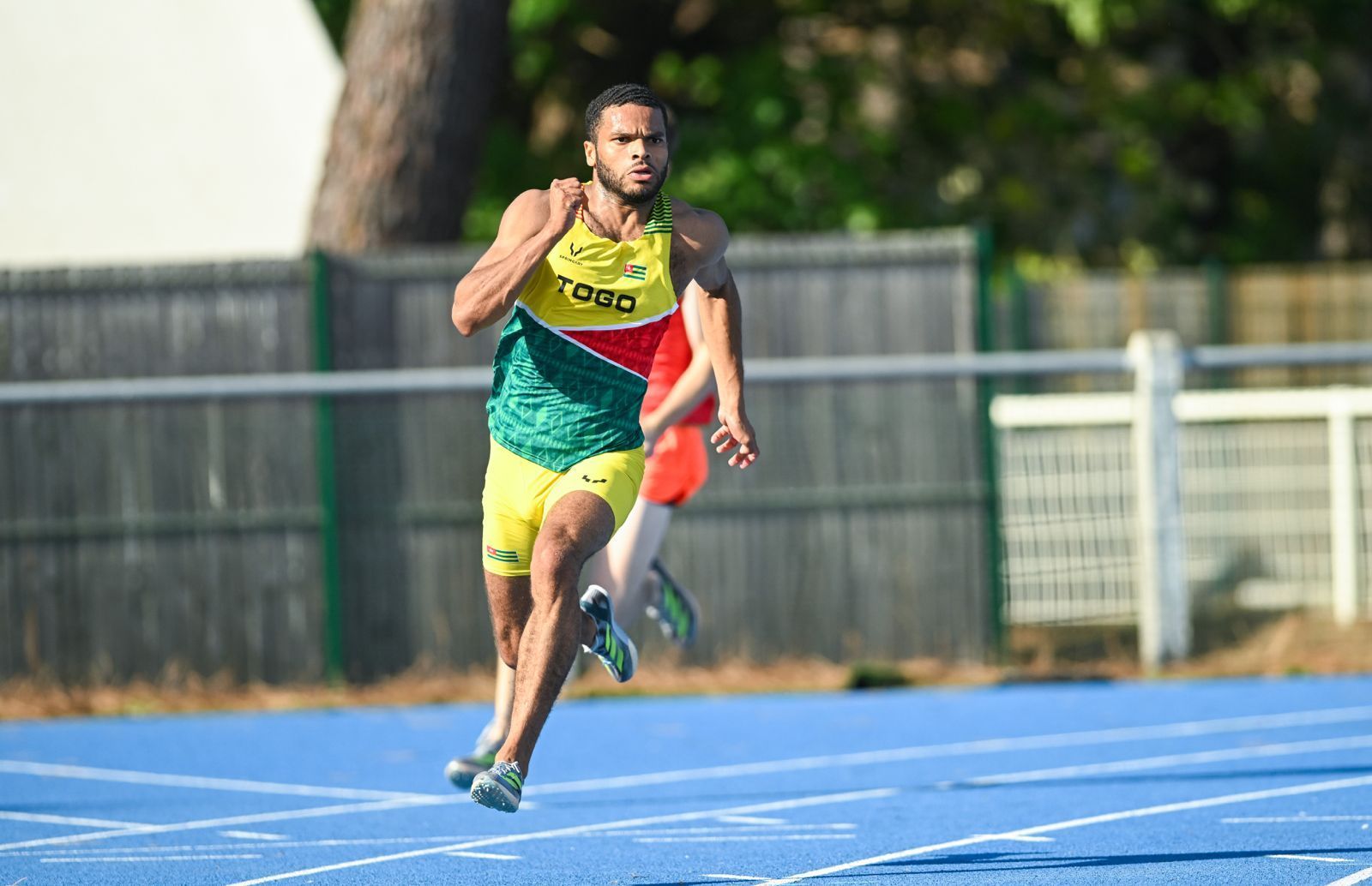 Un athlète en uniforme vert et jaune sprinte sur une piste bleue ; un autre coureur en arrière-plan. Noah Lawson. 
