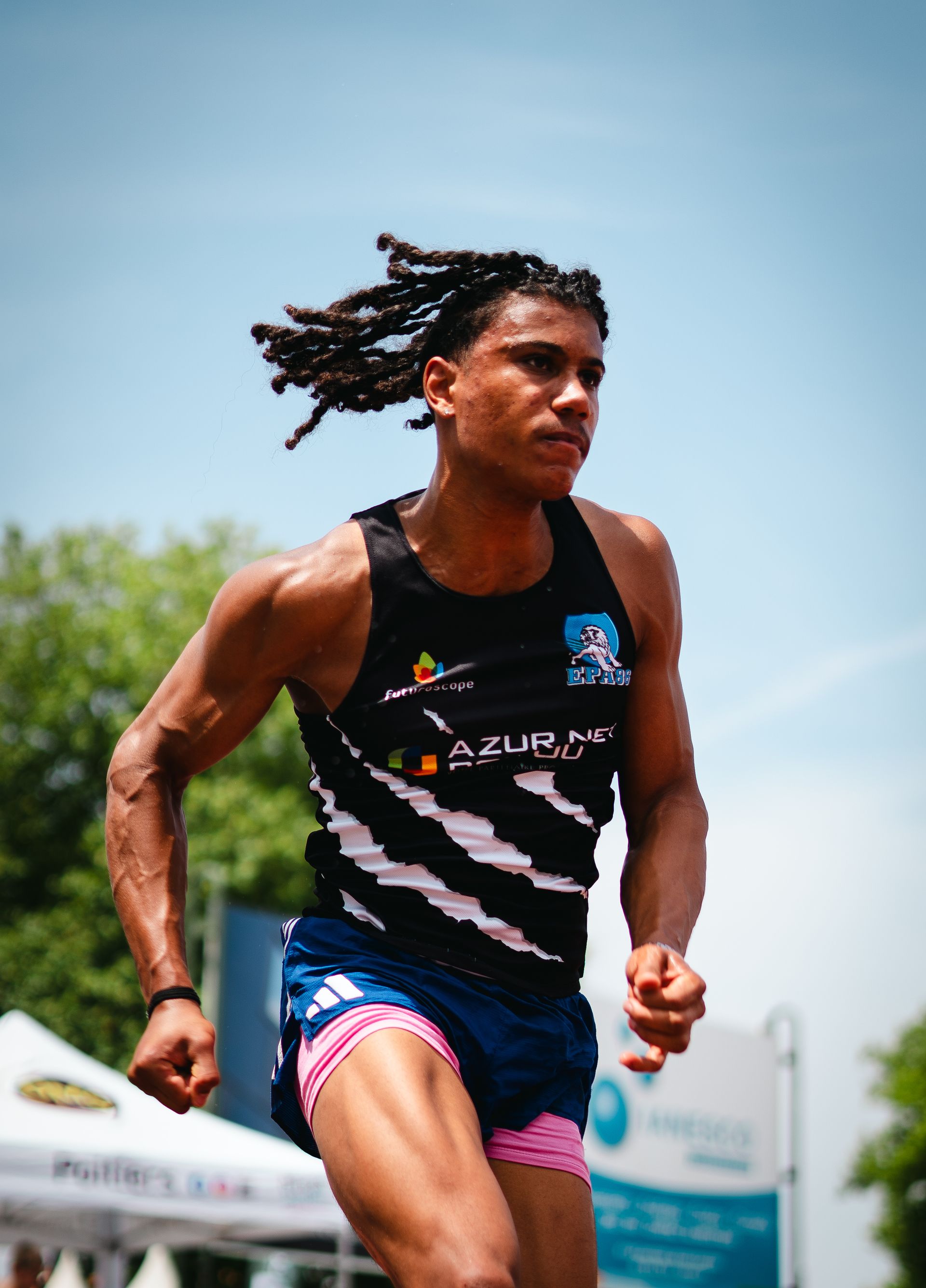 Un jeune athlète court, les cheveux au vent, dans un uniforme bleu et noir ; cadre extérieur ensoleillé. Lucas Blanzac-Joly.
