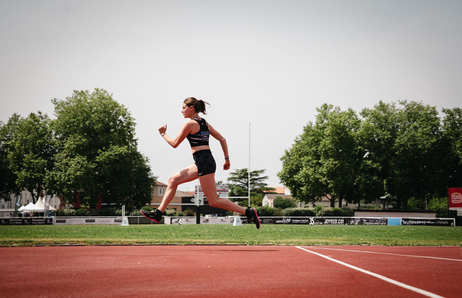 Sauteuse en hauteur prend l'élan sur une piste rouge, sous un ciel ensoleillé, arbres en arrière-plan. Alix Petit. 