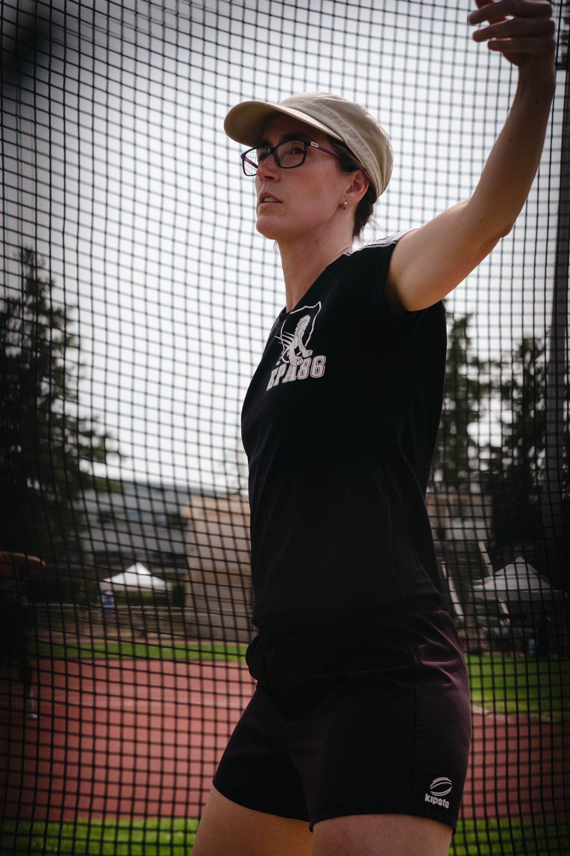 Un athlète vêtue du maillot noir et d'une casquette se prépare à lancer un disque dans la cage de lancer. Mathilde Lambert. 