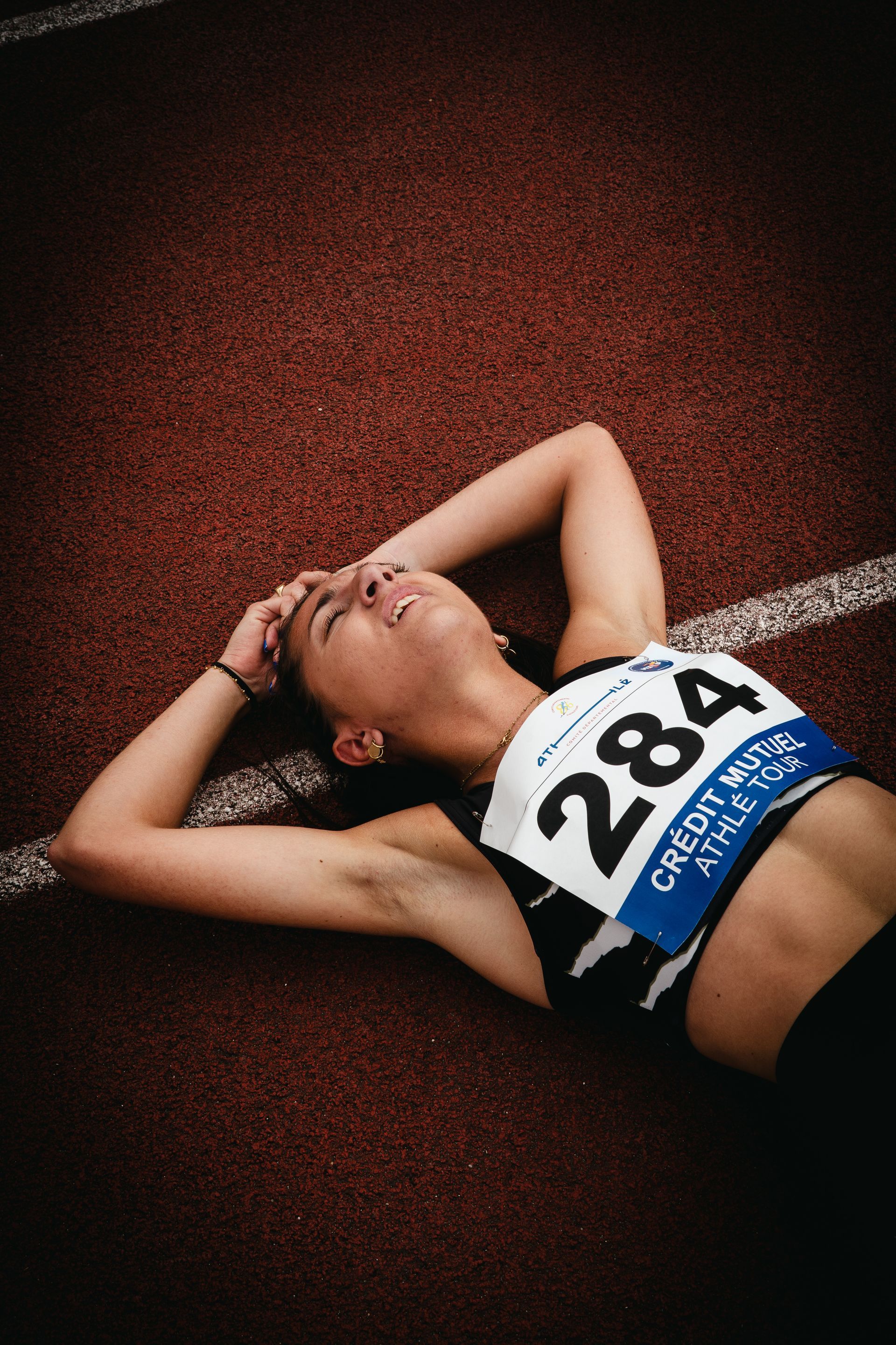 Un athlète épuisé est allongé sur une piste après une course, portant un dossard portant le numéro 284. Melo Chevrier.