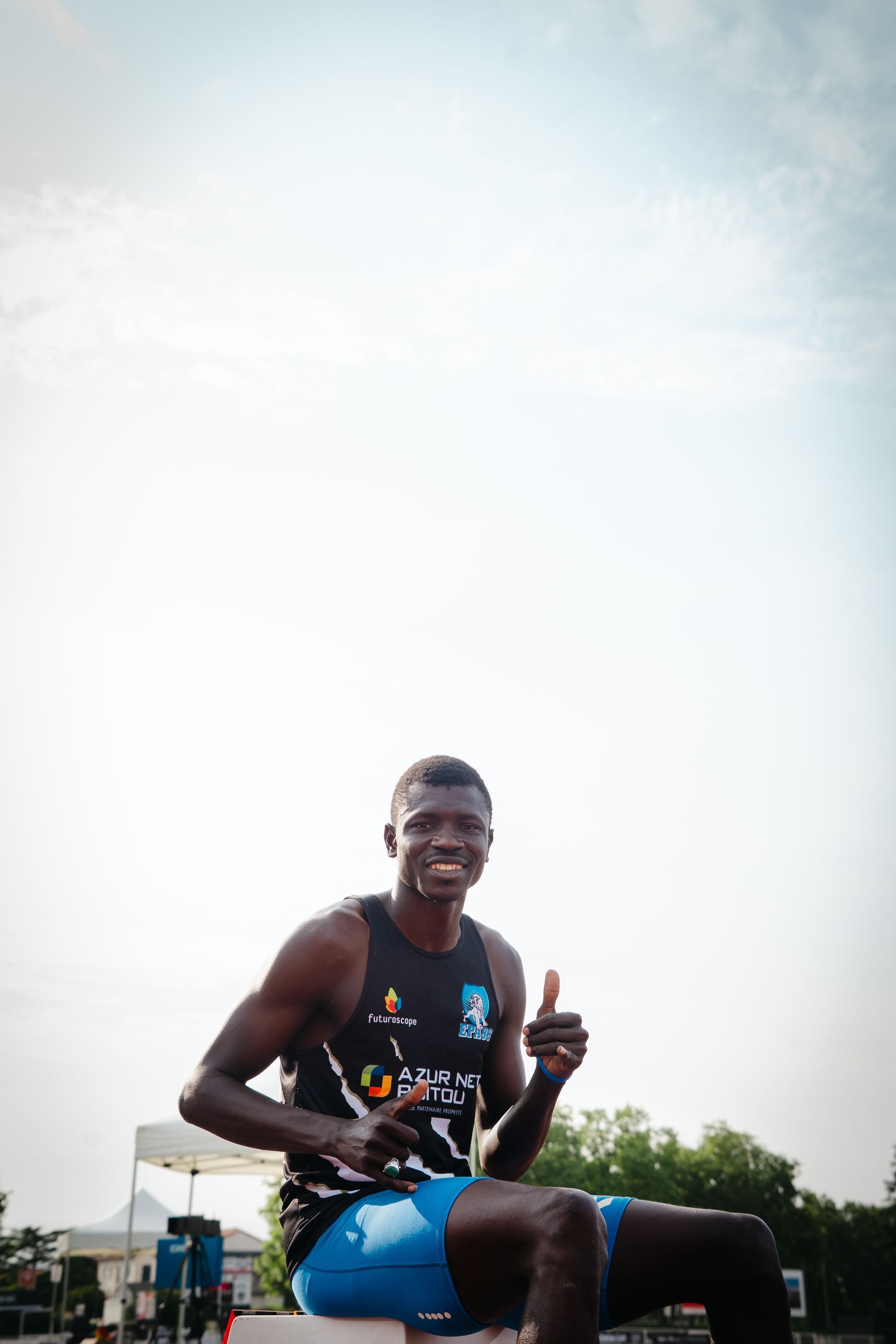 Athlète souriant en débardeur noir et short bleu levant le pouce à l'extérieur.