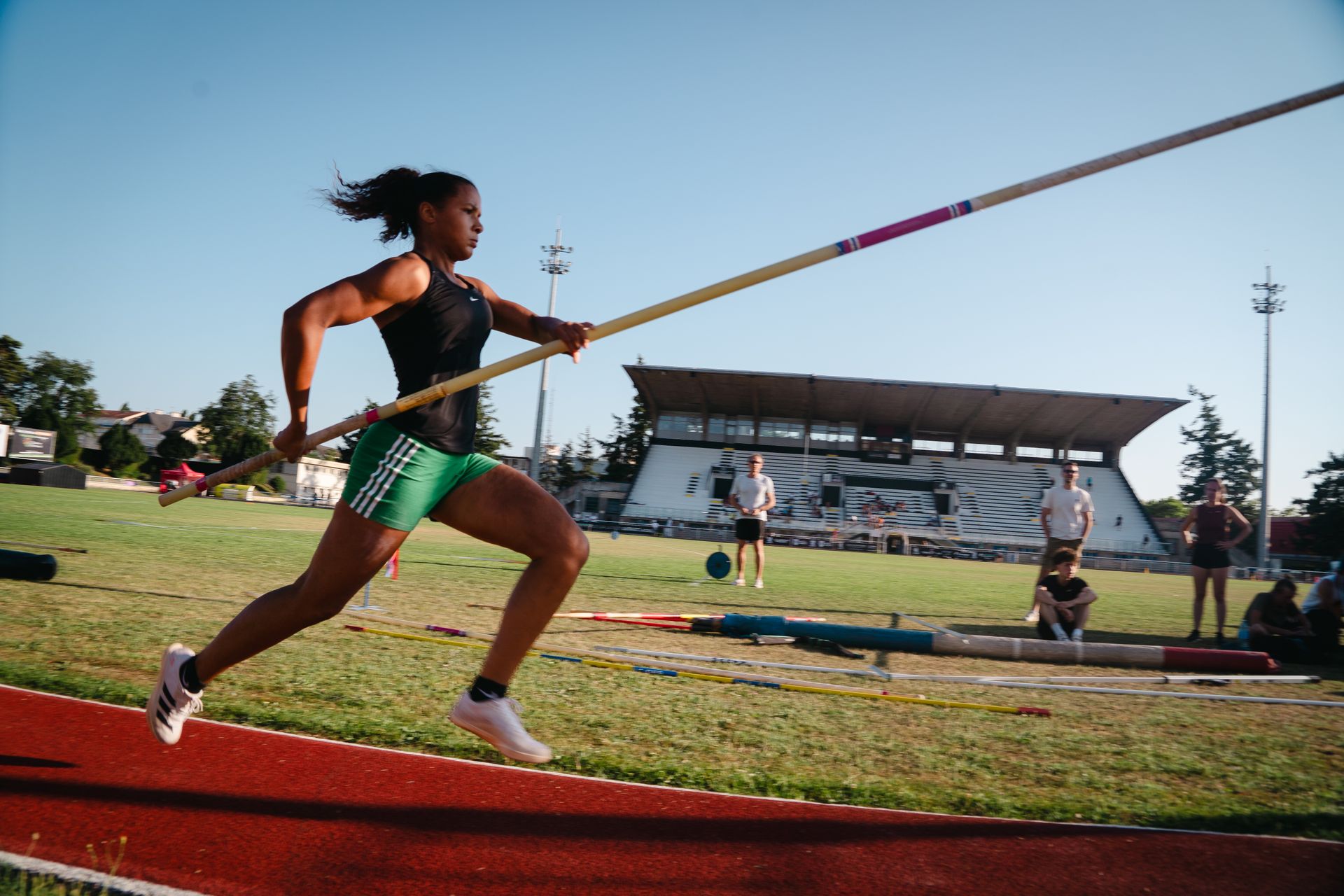 Une perchiste féminine court sur une piste avec une perche à la main, avec un stade en arrière-plan. Neomy Boue. 
