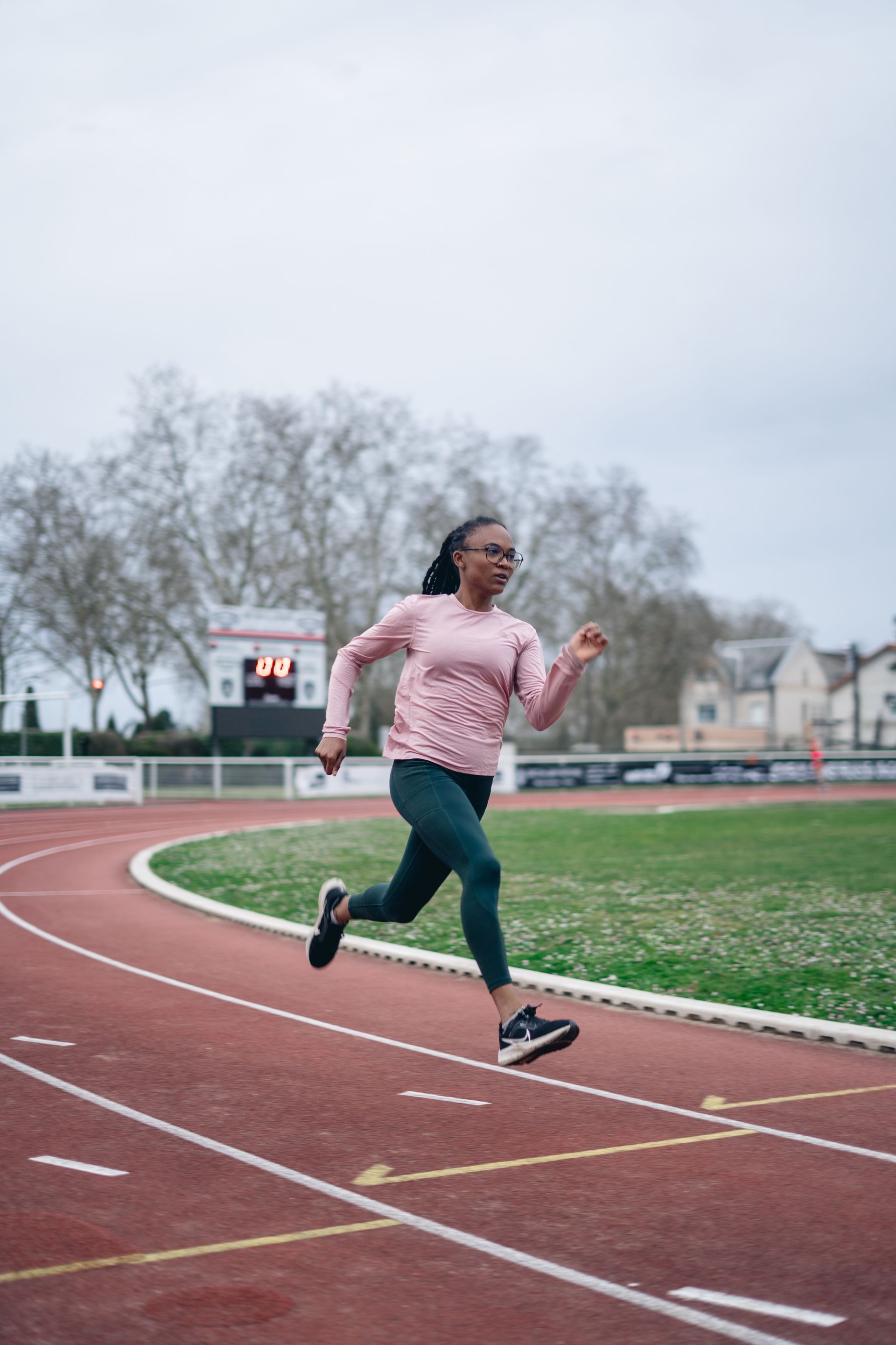 Femme courant sur une piste portant un t-shirt rose et des leggings verts, avec un stade en arrière-plan.