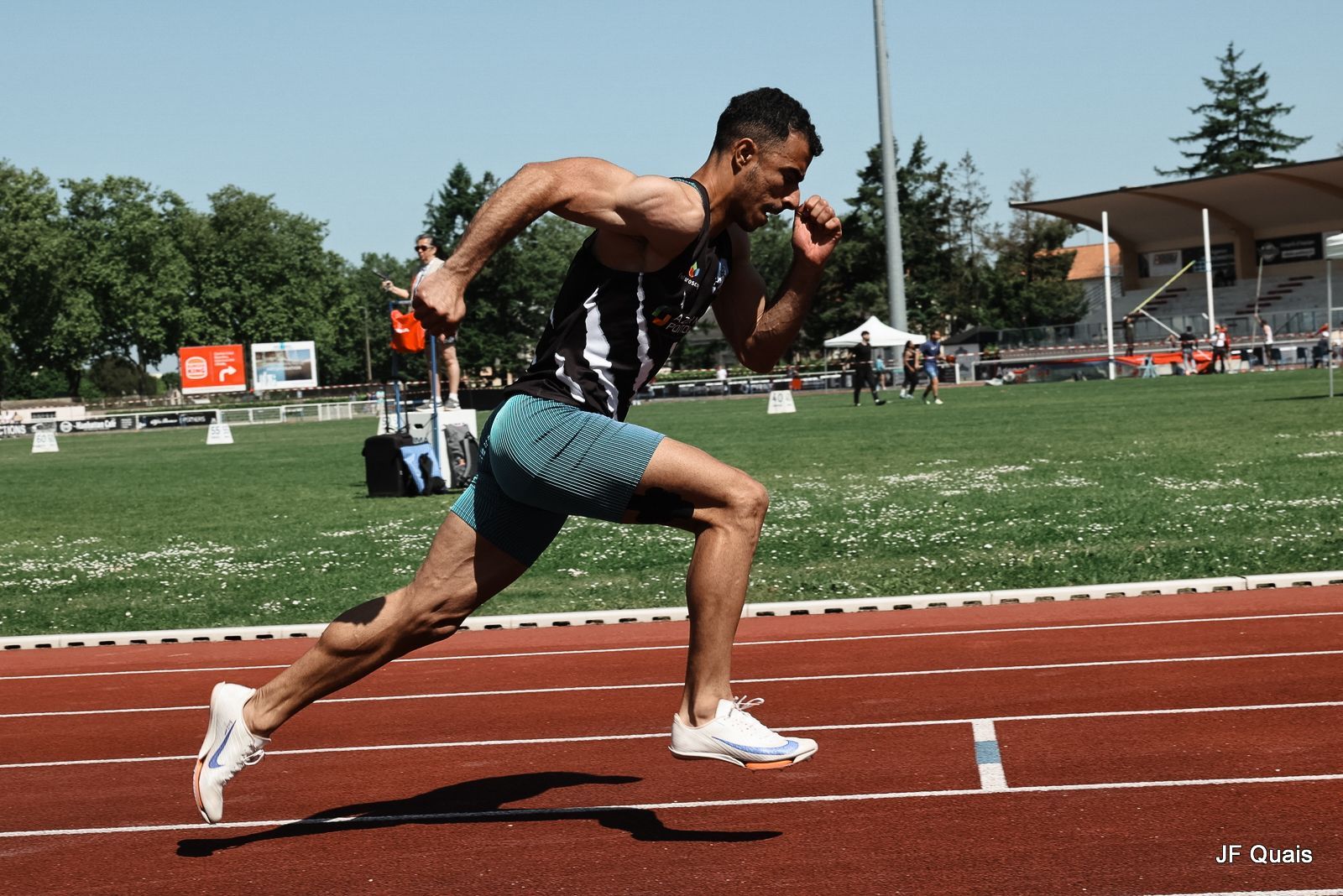 Coureur masculin vêtu d'un débardeur noir et blanc et d'un short bleu sprintant sur une piste rouge. Yayah Nazih