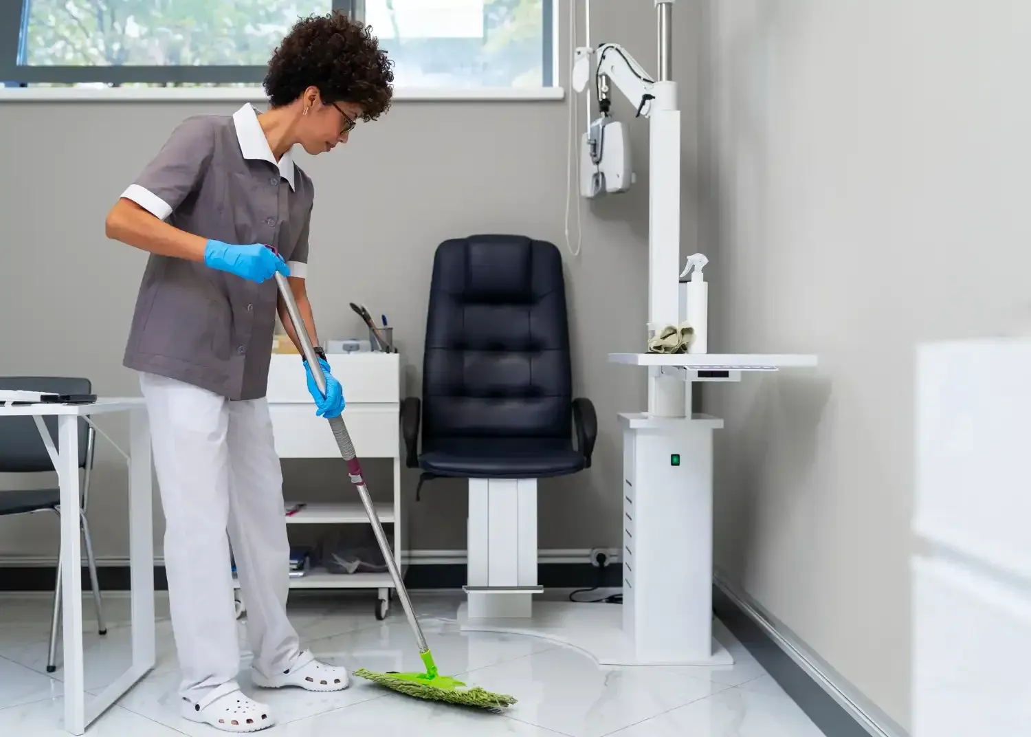 Person mopping a white floor in a clean room with a black chair and window.