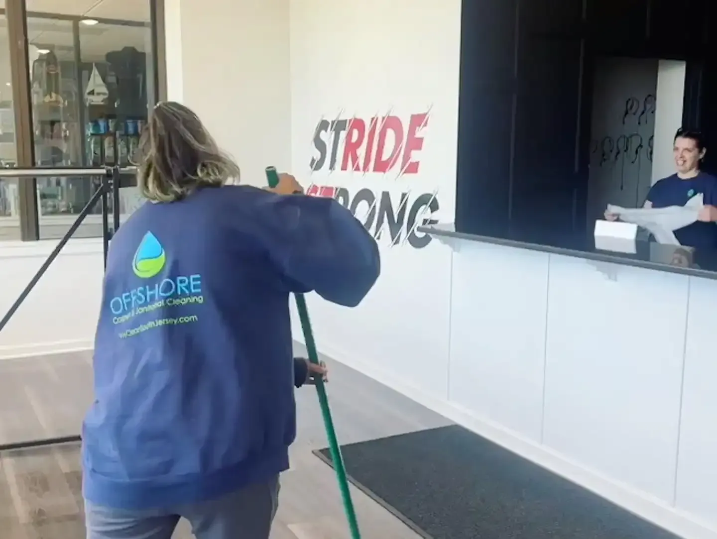 Woman in blue shirt mopping floor at a business, another woman behind a counter. 