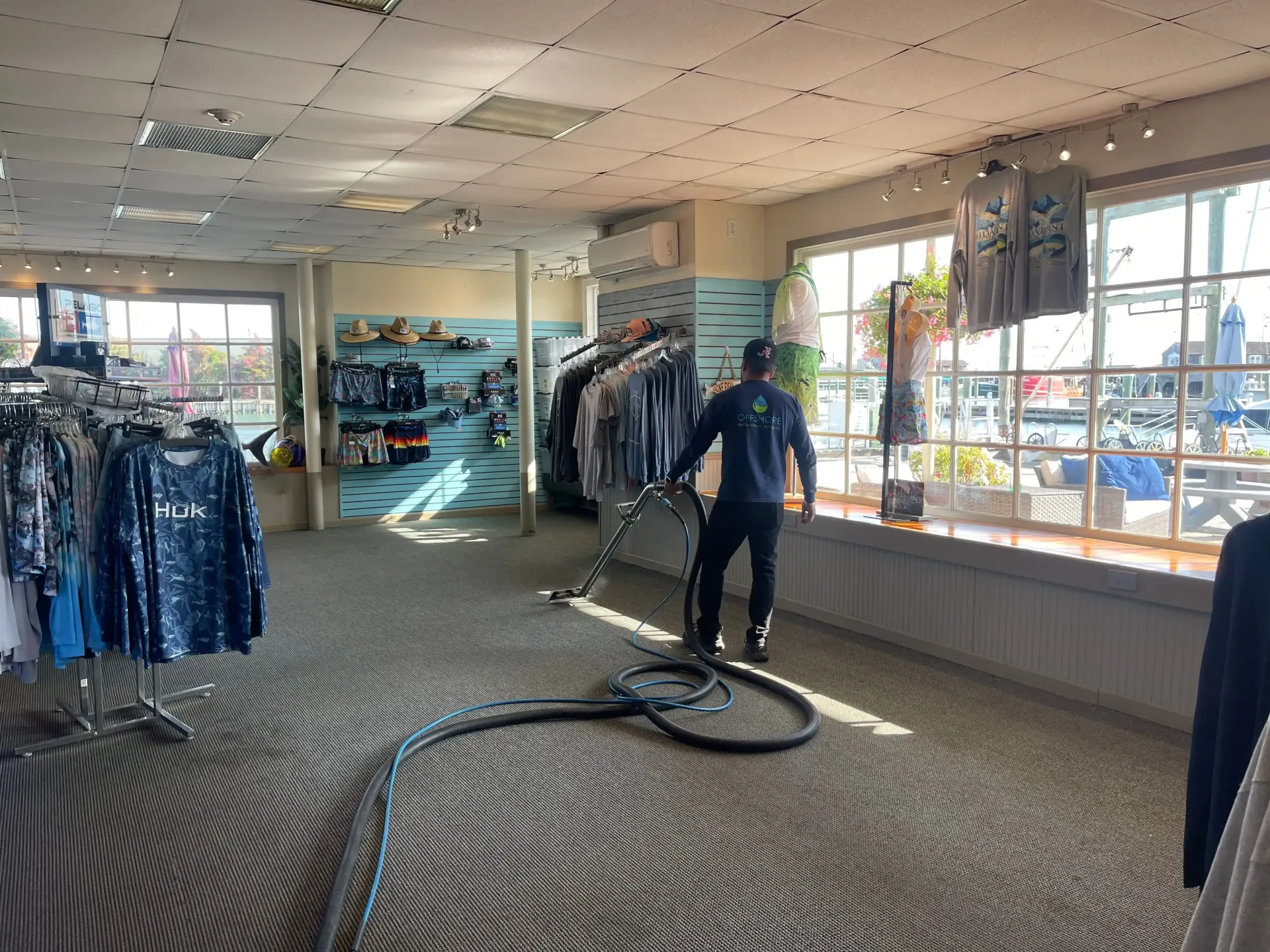 Person vacuuming the carpet of a clothing store with racks of clothing and large windows.