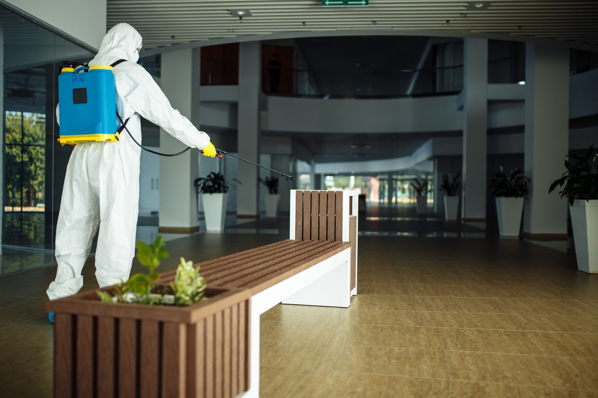 A man in a protective suit is spraying disinfectant on a bench.