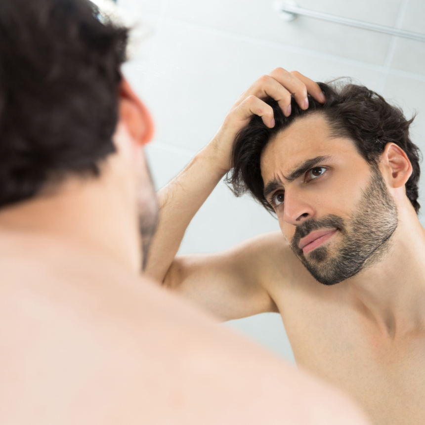 Man looking in mirror, touching hair, concerned expression. Bathroom setting, white tile.