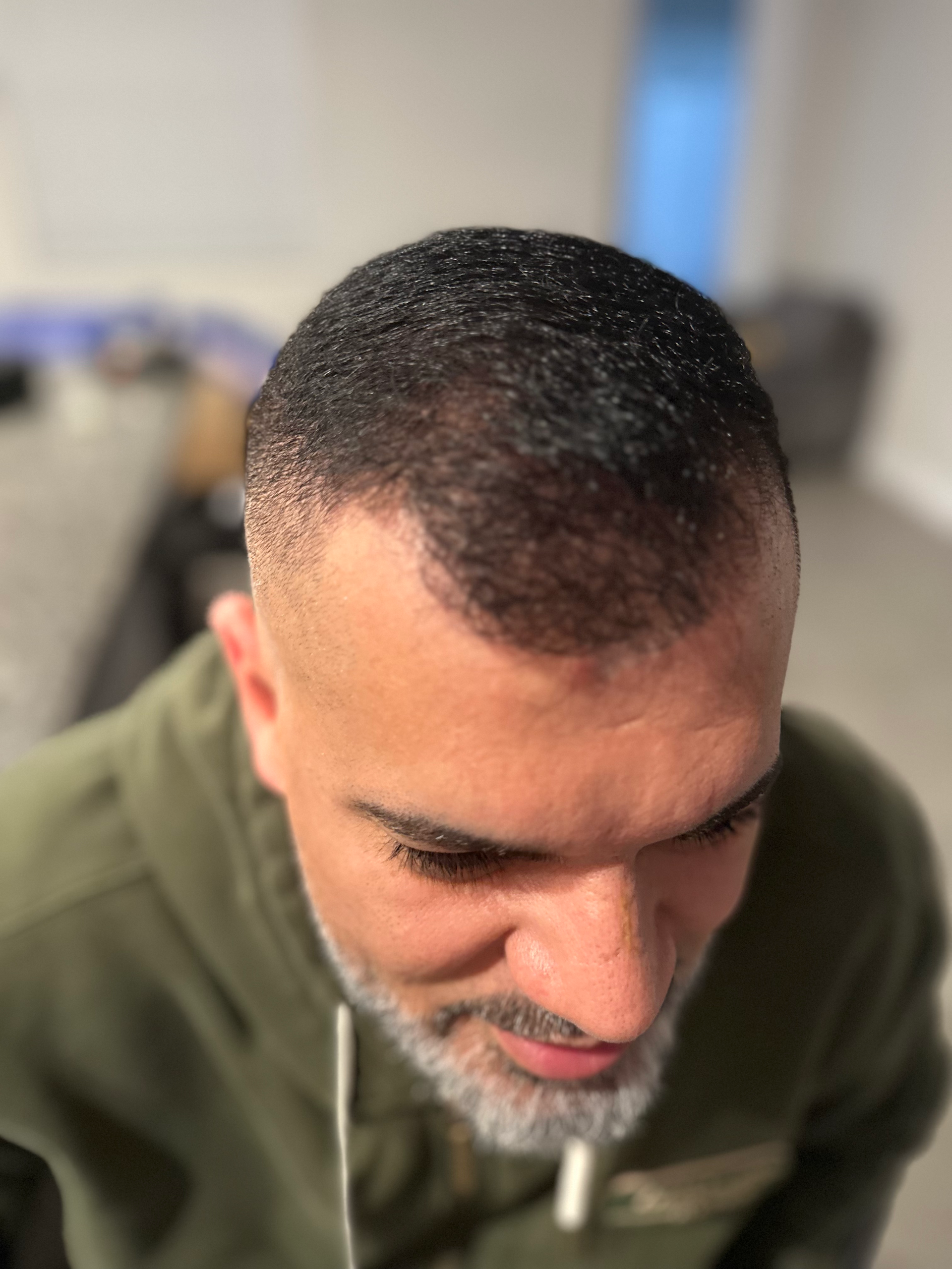 Man with shaved head and beard, wearing a green hoodie, looking down, indoors.