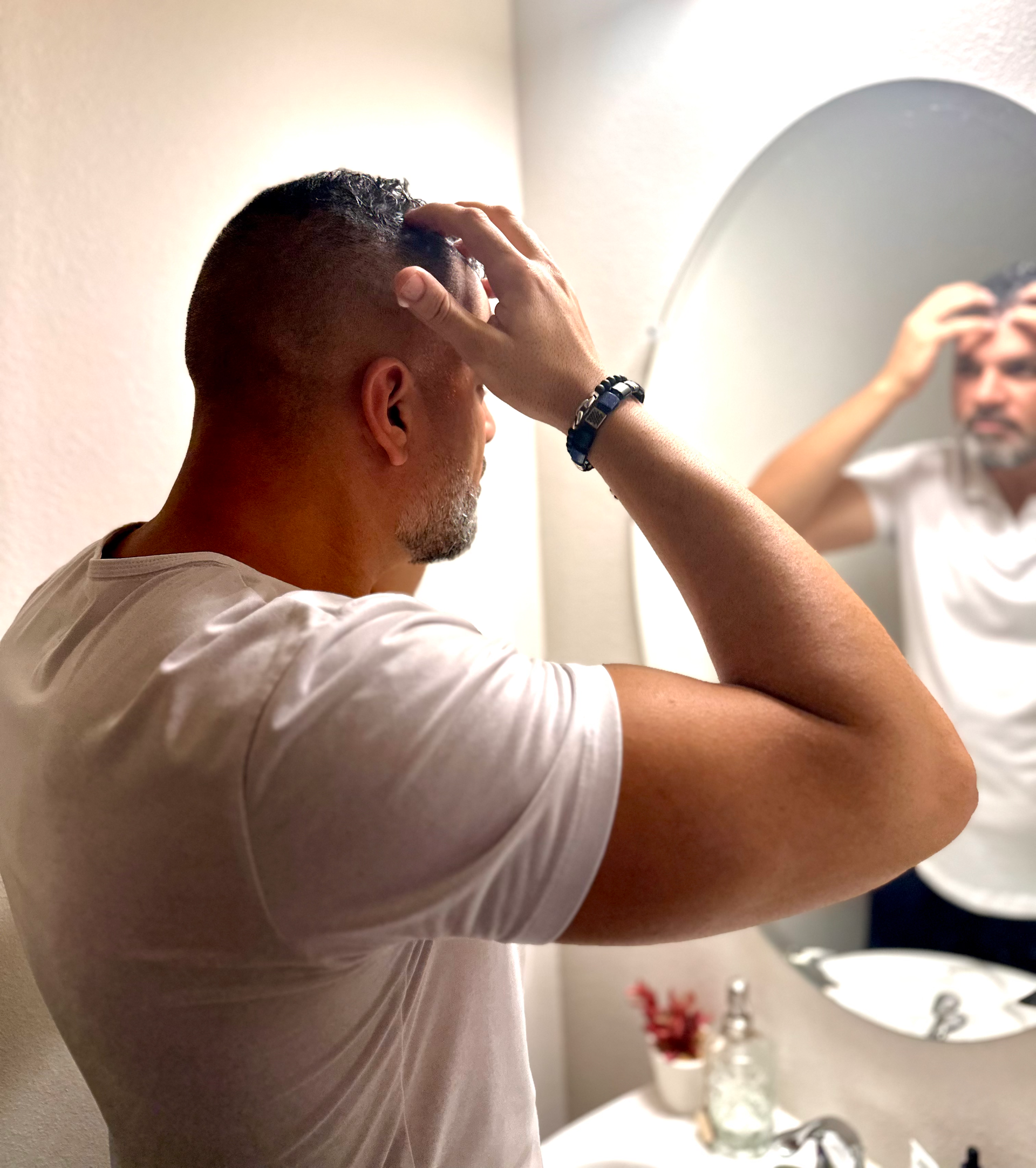 Man looking at his hair in a bathroom mirror, white shirt, touching his head.