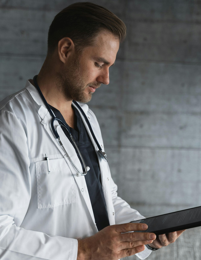Doctor in white coat, stethoscope, looking down at tablet, against a concrete wall.