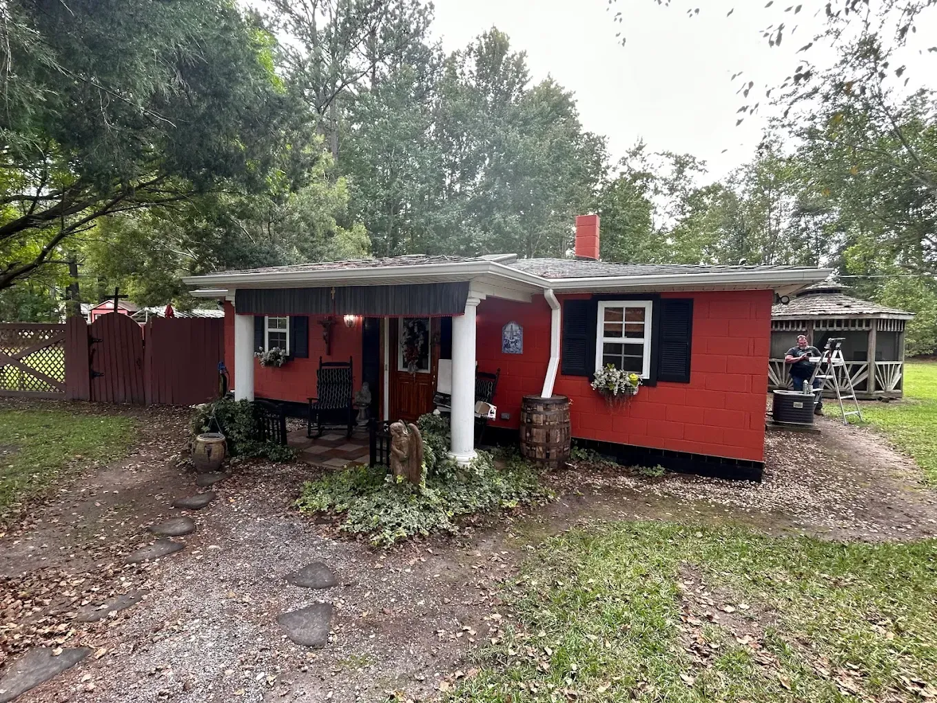 Red cottage with white columns, black shutters, and a small gazebo in the background.