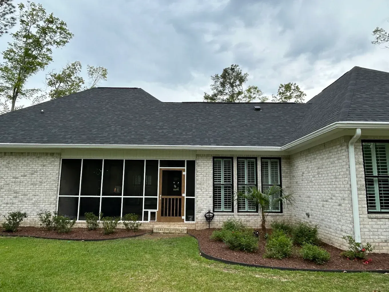 Backyard view of a house with a dark roof, screened-in porch, brick exterior, and lush landscaping.