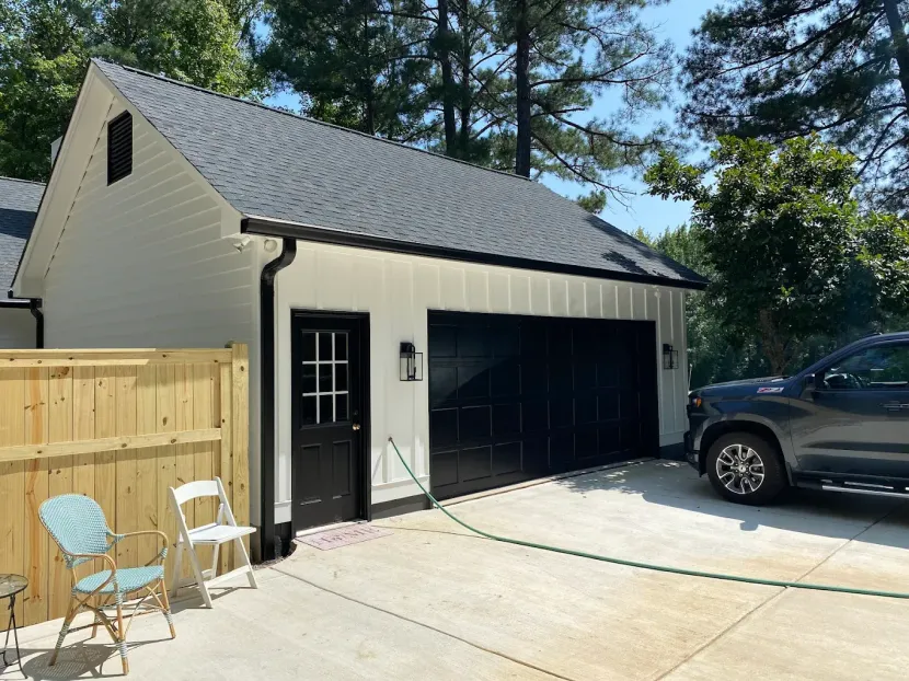 White-sided detached garage with black trim, door, and garage door. A truck is parked to the right. A small fence and chairs are in front.