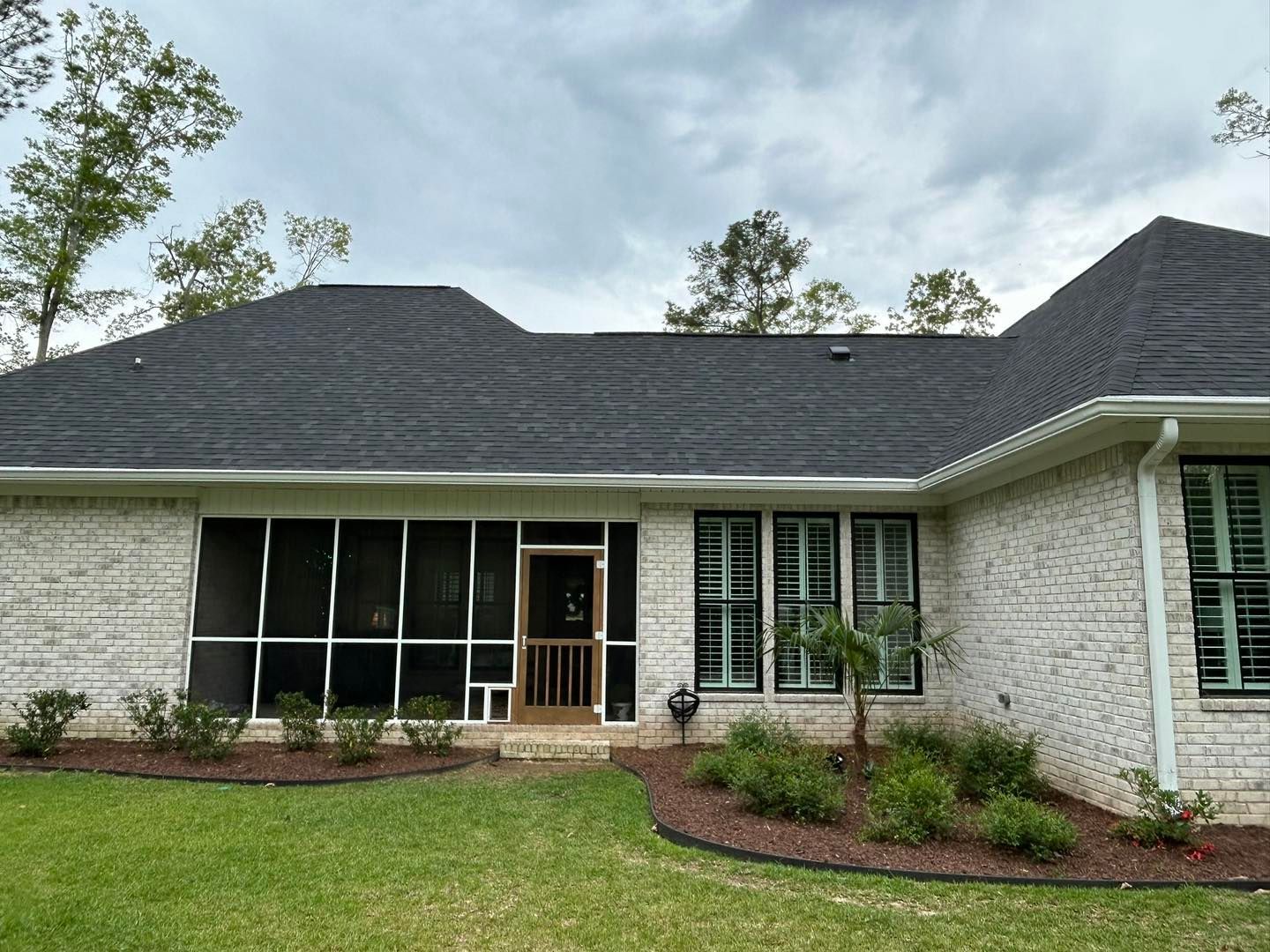 Exterior of a light brick house with a dark roof and a screened porch, surrounded by green grass and landscaping.