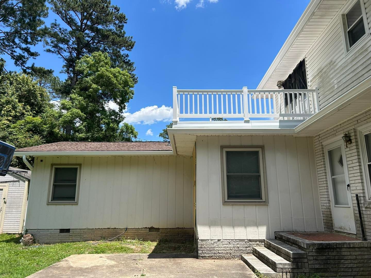 Exterior of a white two-story house with a deck. Blue sky, green trees, and concrete patio.