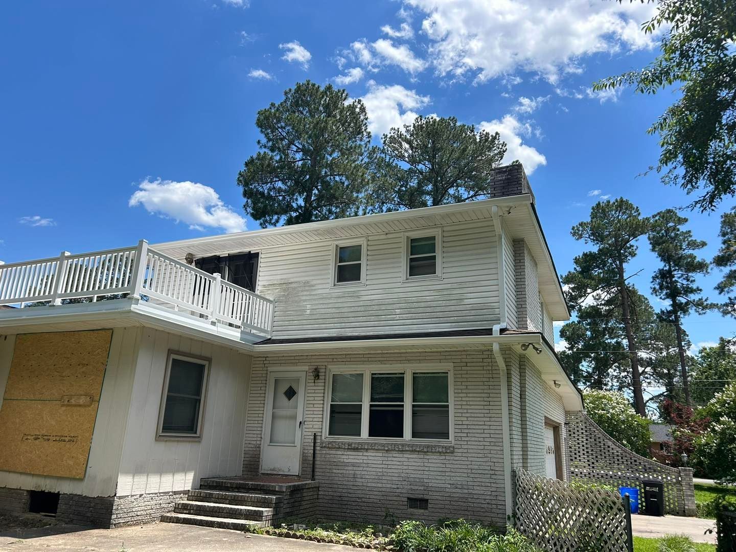 Two-story white house with boarded-up windows and a balcony, under a cloudy blue sky.