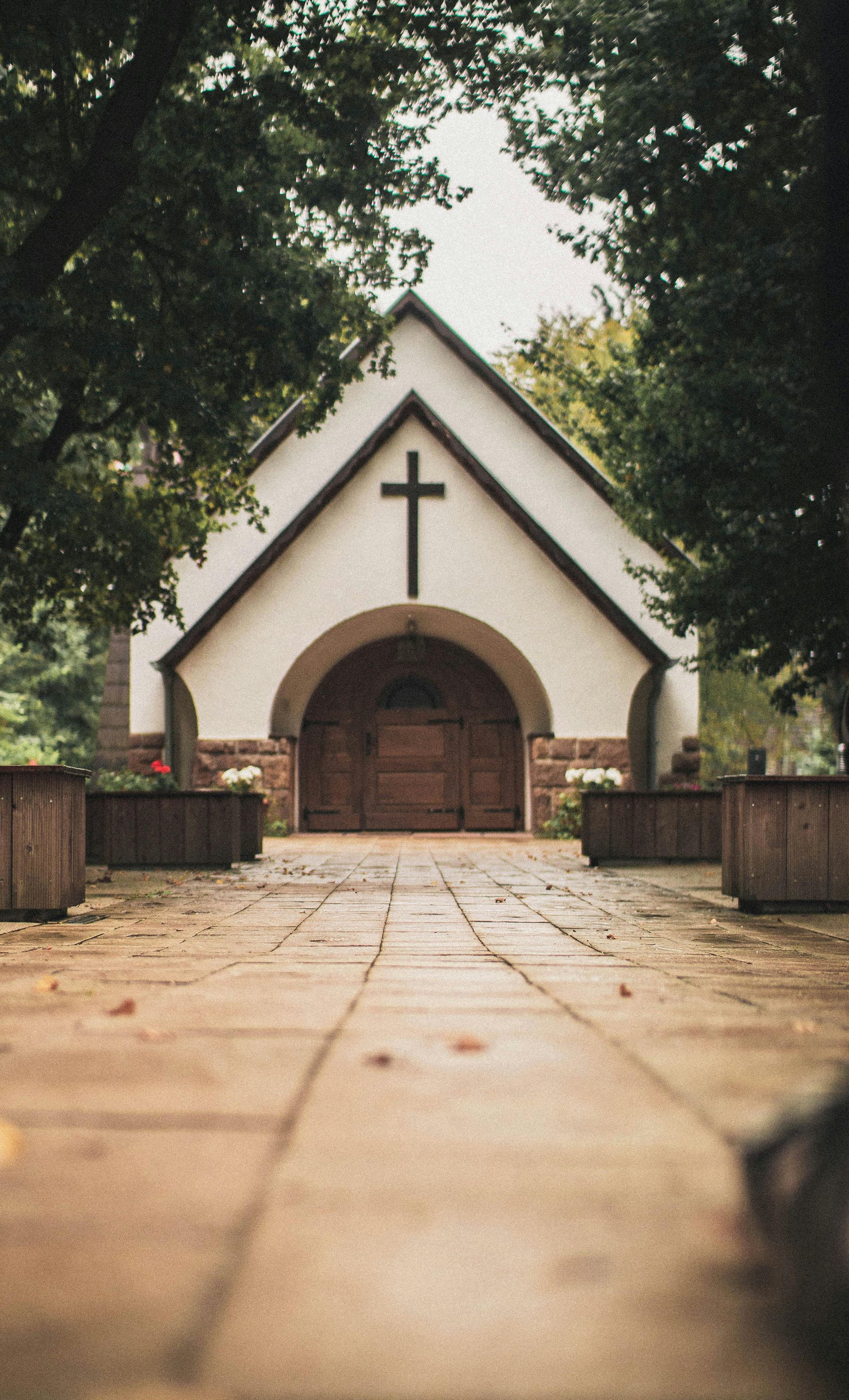 Chapel with cross on white facade, brown arched entrance, stone walkway, trees in background.