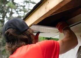 Person in red shirt and cap installing soffit panel under roof overhang.