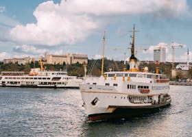 White and yellow passenger ferry sailing on a waterway, with buildings and blue sky in the background.