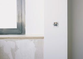 White wall with a window, outlet, and unfinished plaster.